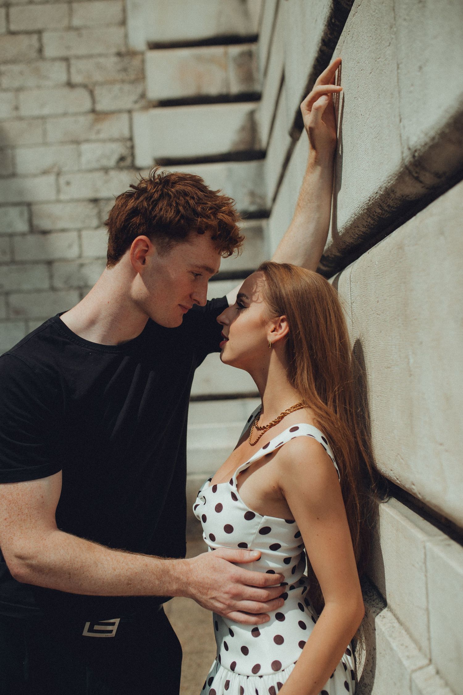 A romantic couple shares an intimate moment against a white stone wall on a sunny day.