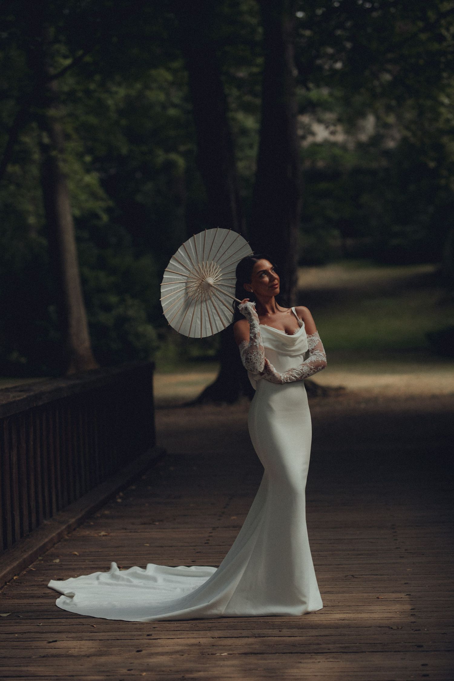 Bride in white satin gown with lace sleeves holds paper parasol on wooden bridge surrounded by lush green forest.