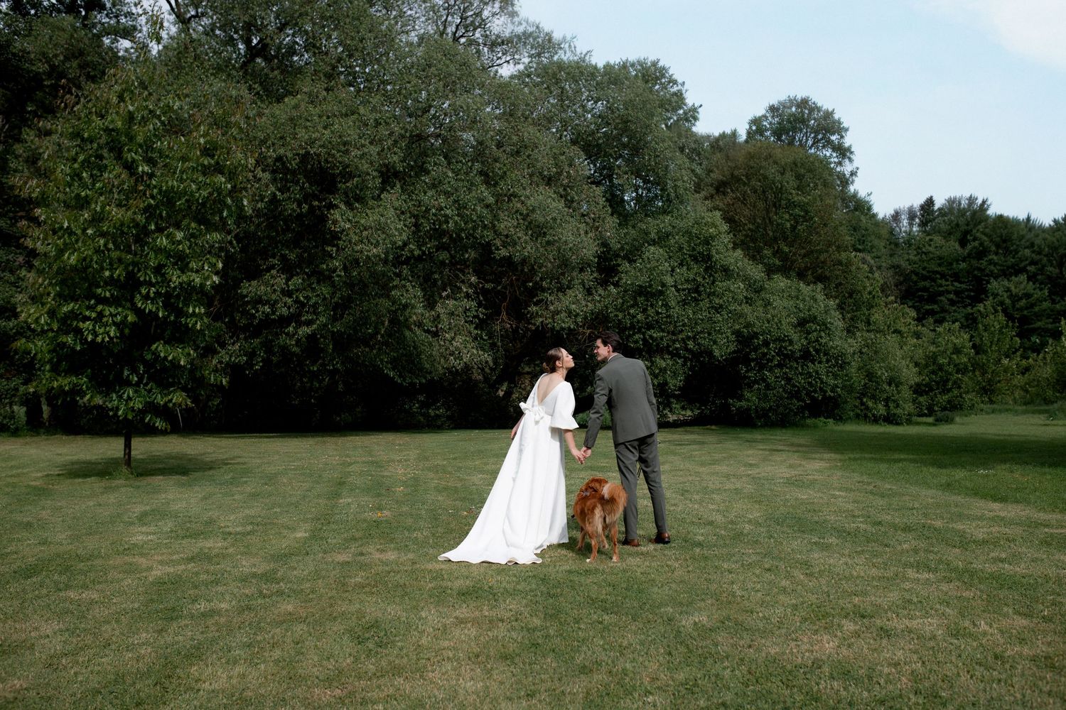 Bride and groom standing with their dog on a lush green lawn surrounded by tall trees on their wedding day.
