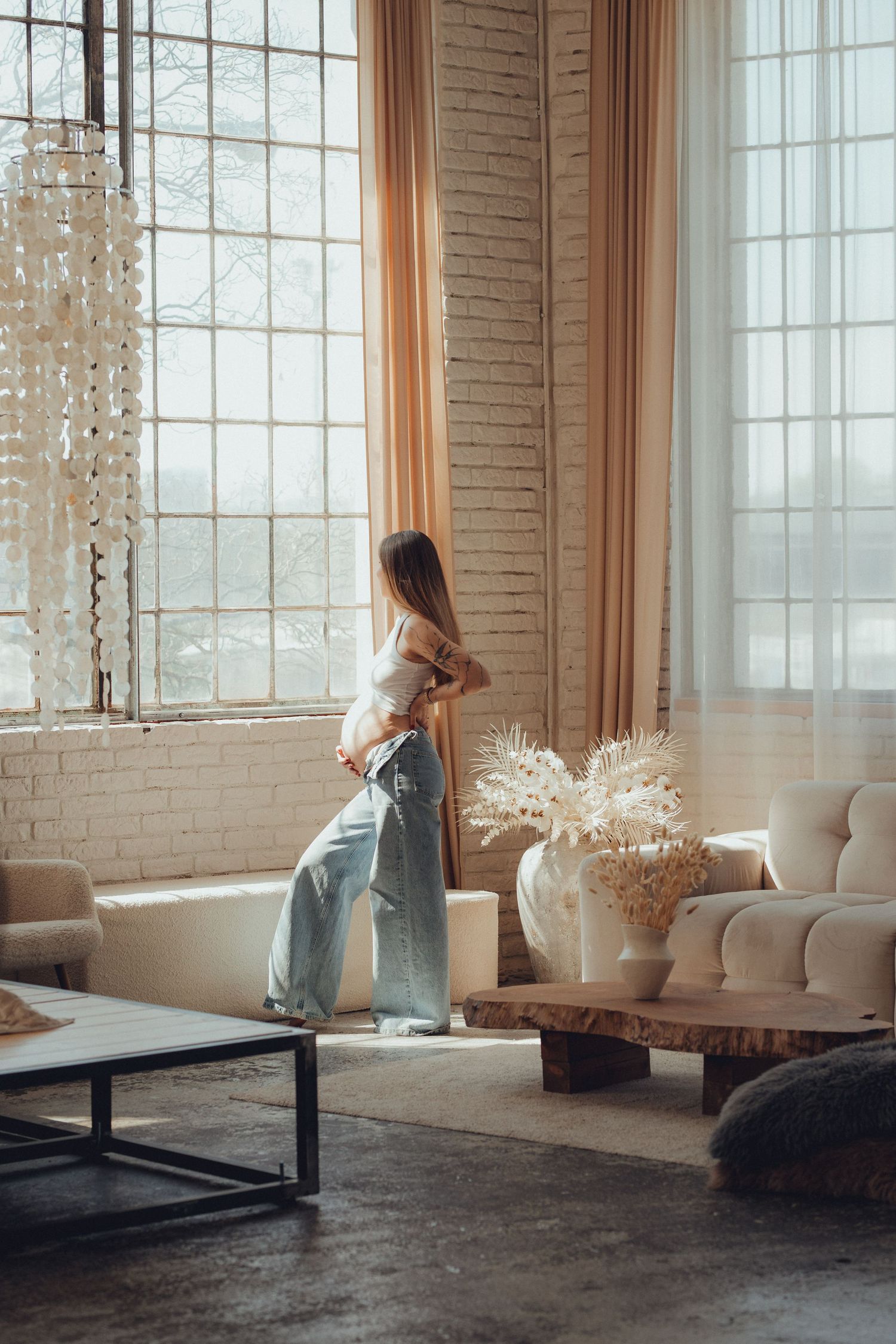 Expectant mother in jeans and white top standing by large windows in modern room with soft natural lighting.