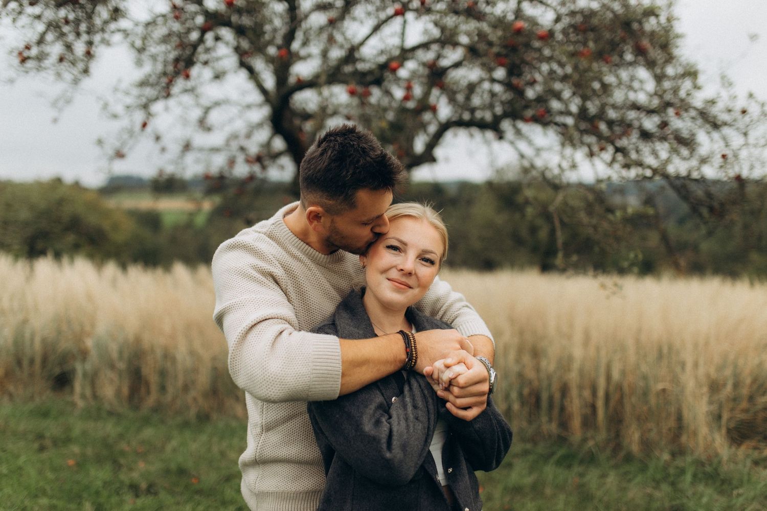A couple embraces in a sunlit field surrounded by autumn trees and tall grass.