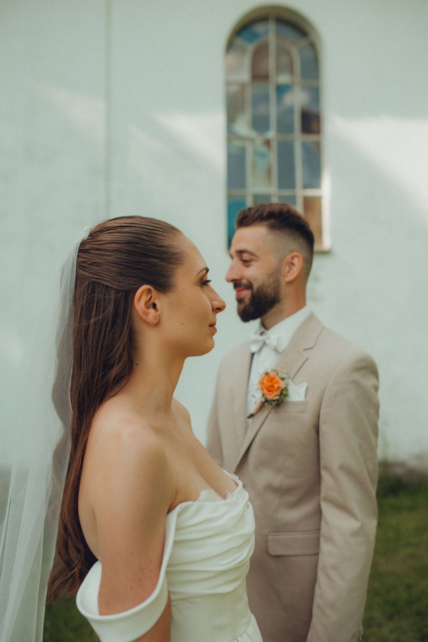 A bride in a white off-shoulder dress and groom in beige suit share tender moment outside a church with arched window.