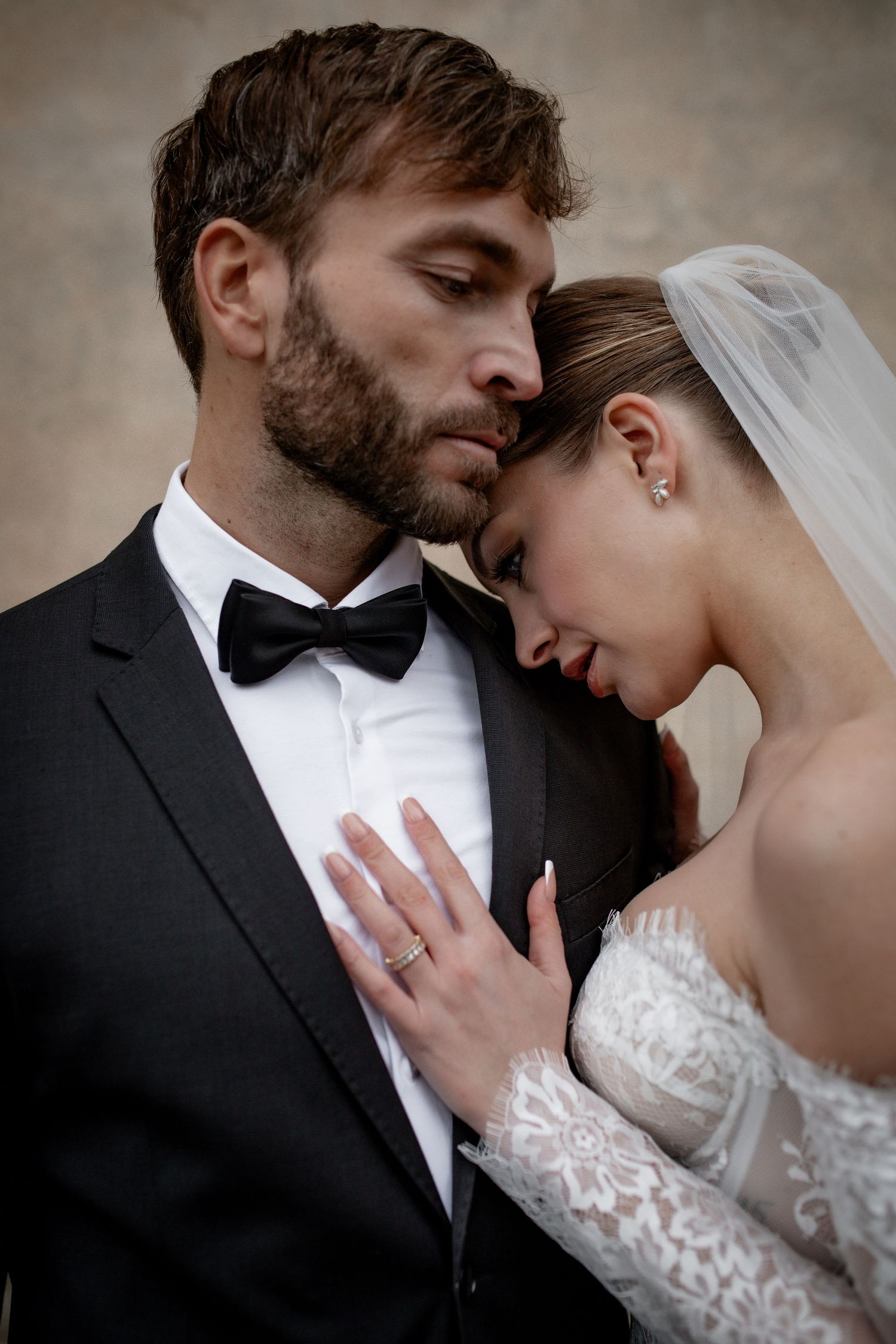 Bride in lace gown and veil leans against groom in black tuxedo and bow tie in an intimate wedding portrait.