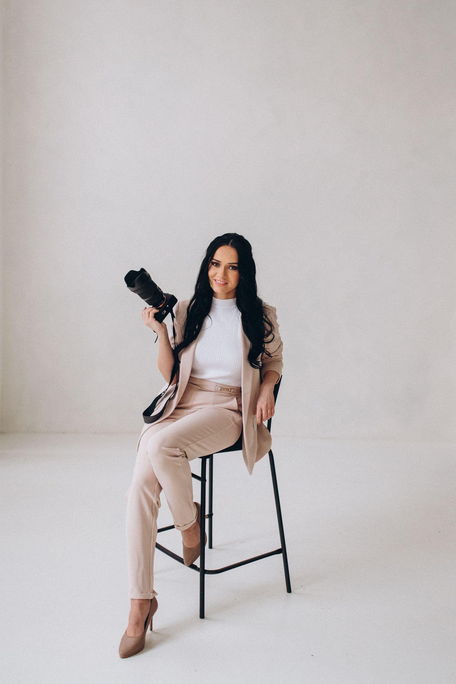 Professional photographer in beige outfit posing with camera on minimalist stool.