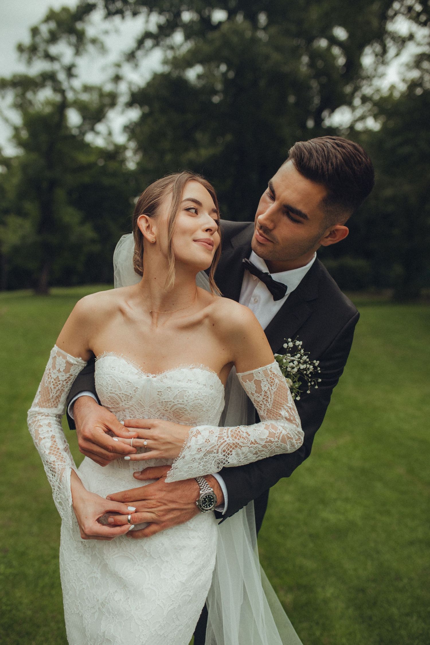 Bride and groom embrace in a lush green park, she wears a lace off-shoulder gown as he holds her in a black tuxedo.