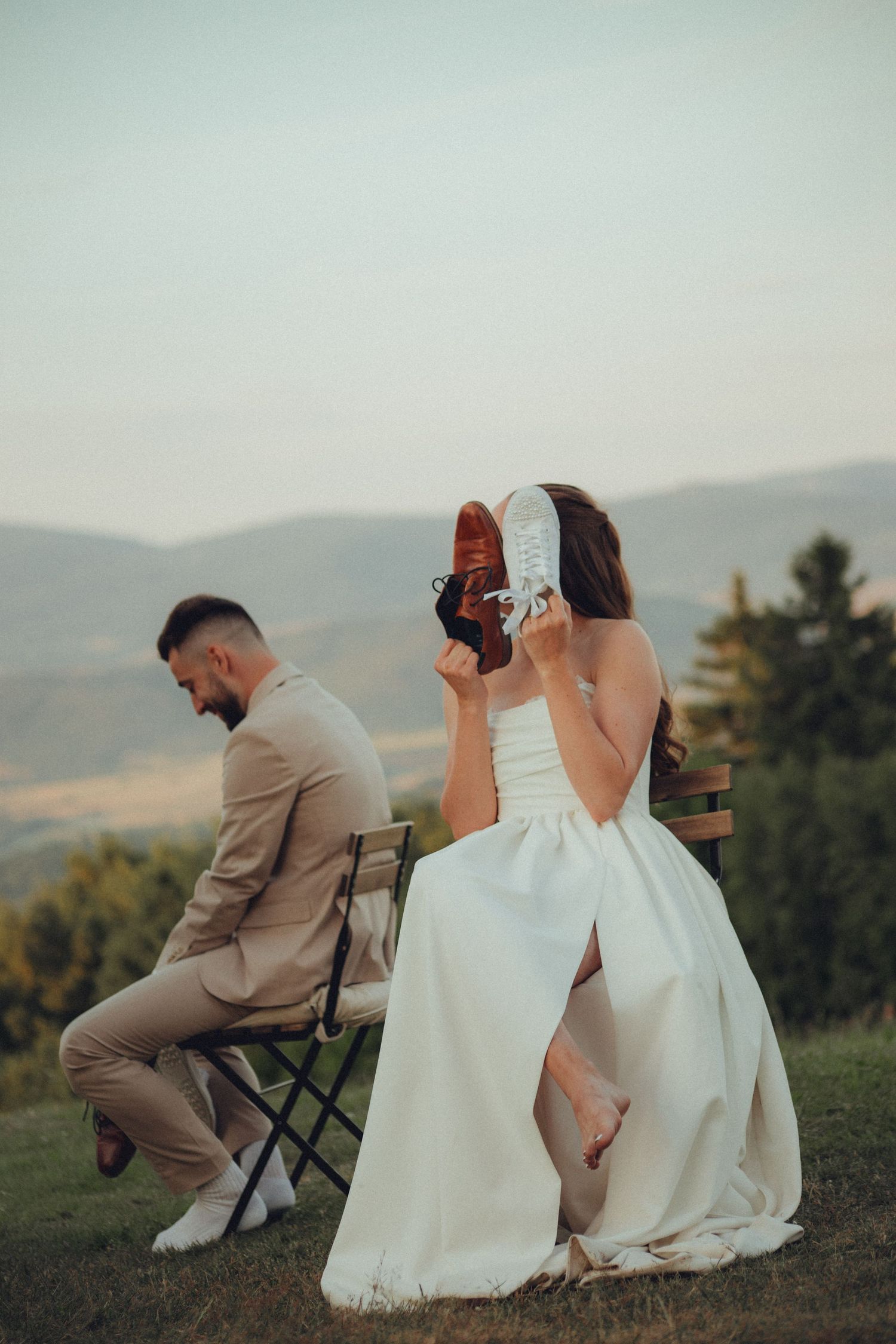 A couple in wedding attire sits together on a hillside during sunset overlooking scenic mountain views.