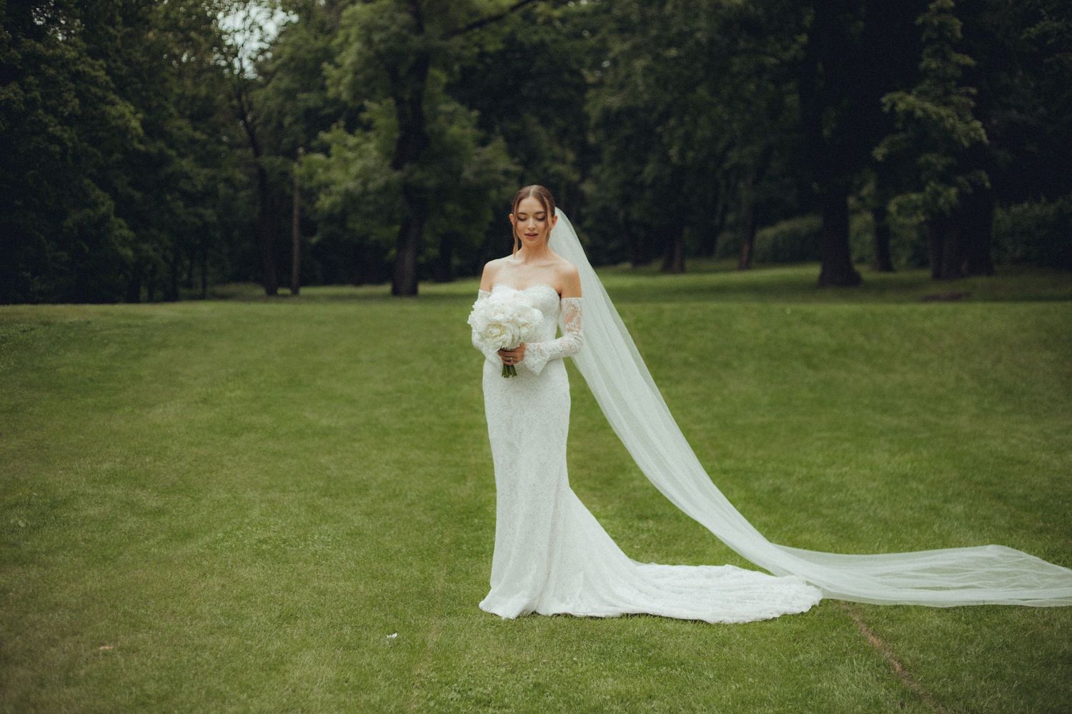 Bride in lace gown with flowing veil holds white bouquet on green lawn.