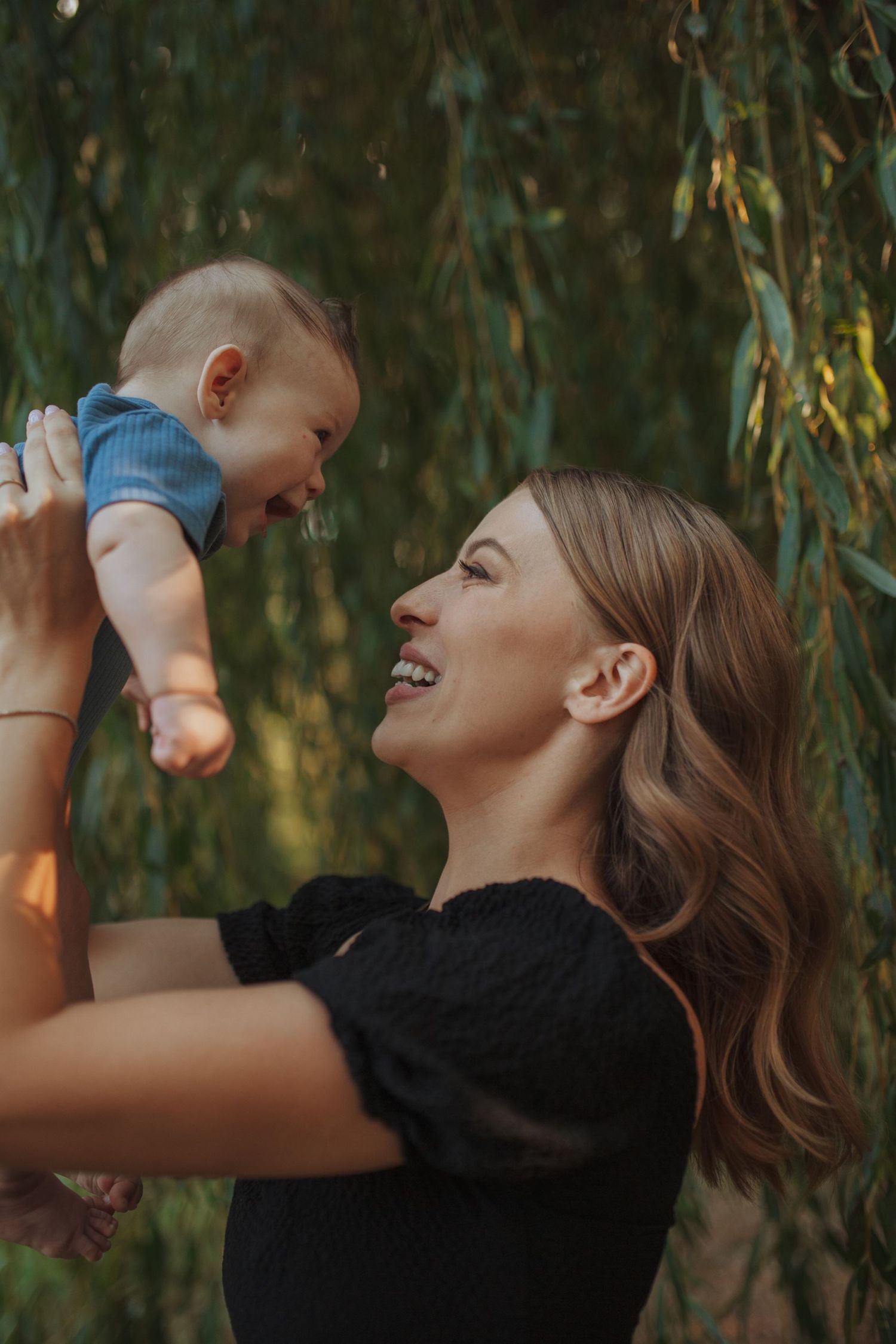 Parent lifting baby in air while laughing under willow tree branches.