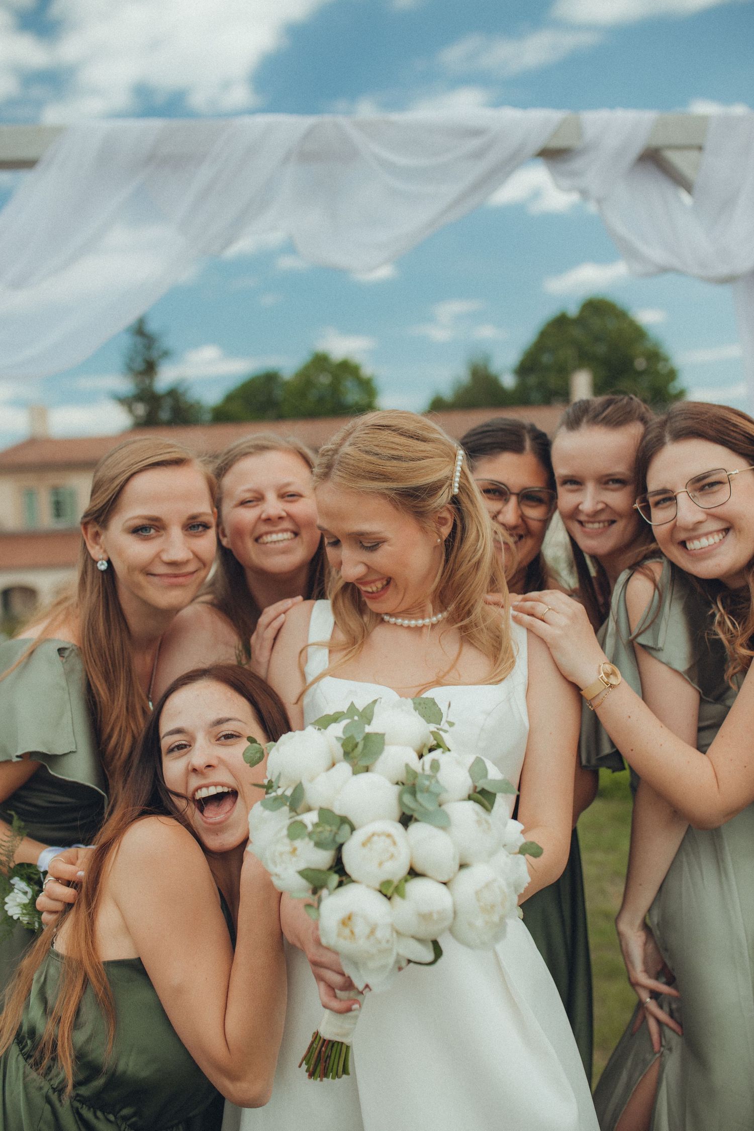 Bridal party in sage green dresses sharing laughs with white floral bouquets.