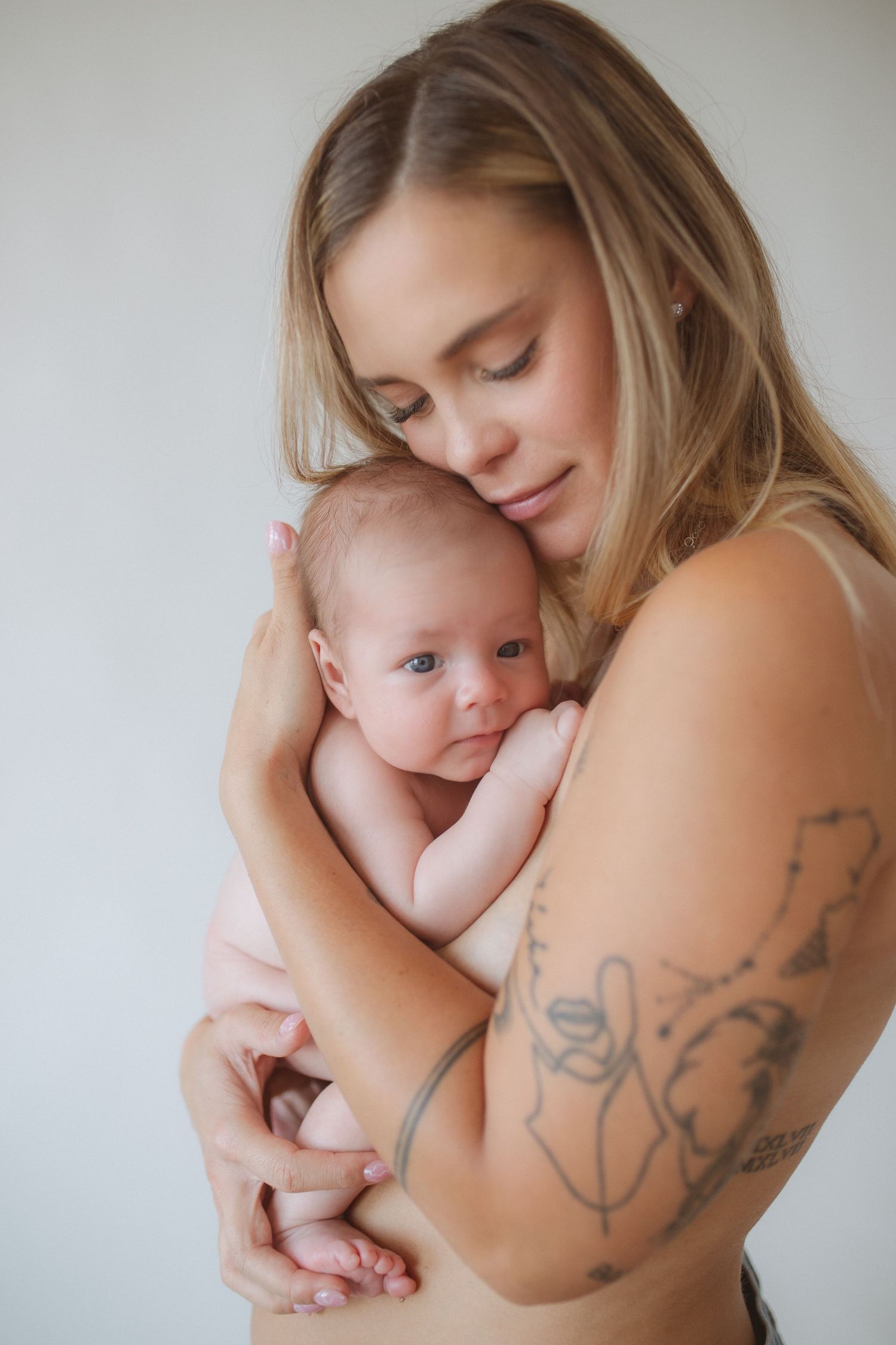 Intimate motherhood portrait with baby against white background showing artistic tattoos.