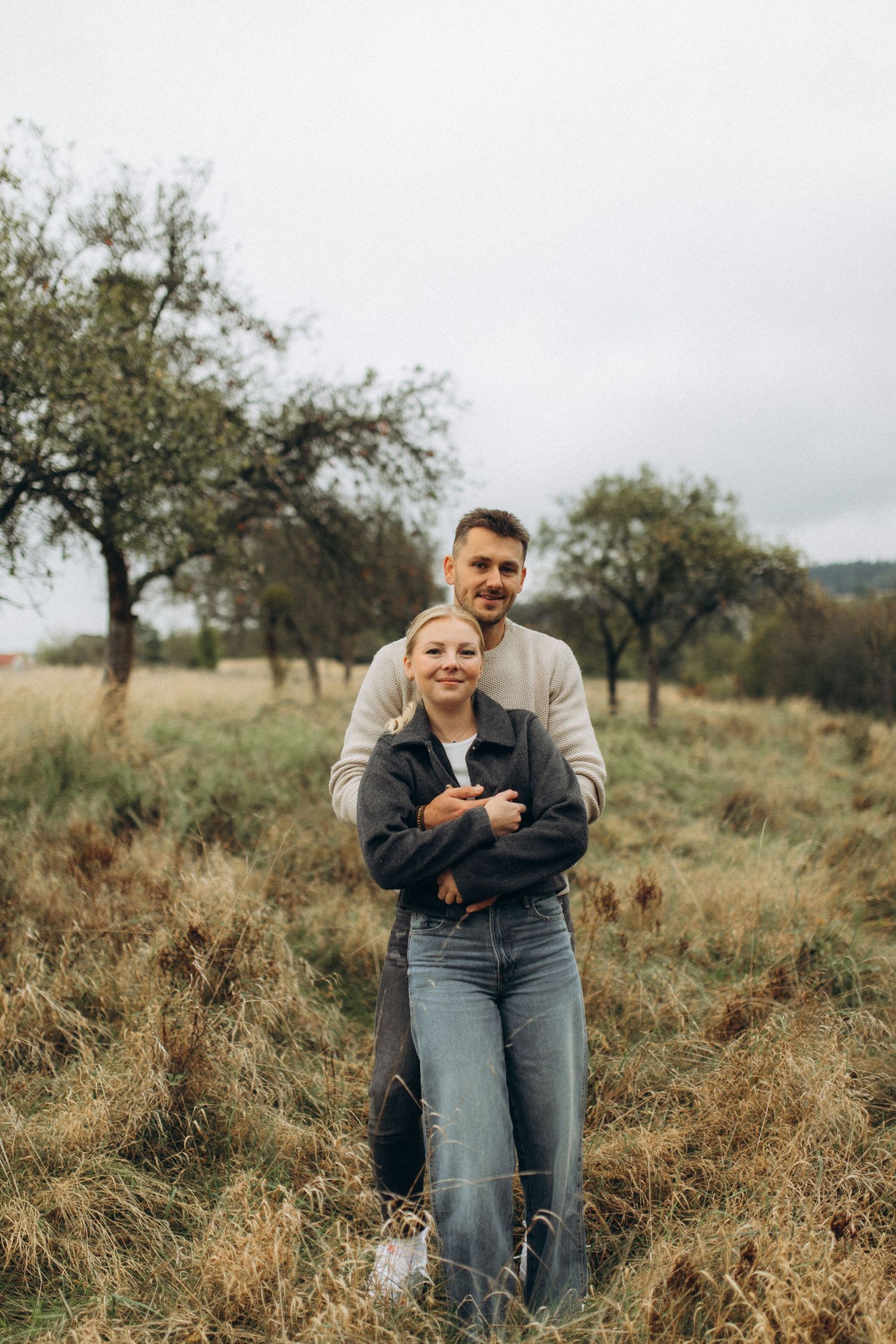 A couple embraces while standing in a field with scattered trees on an overcast day.