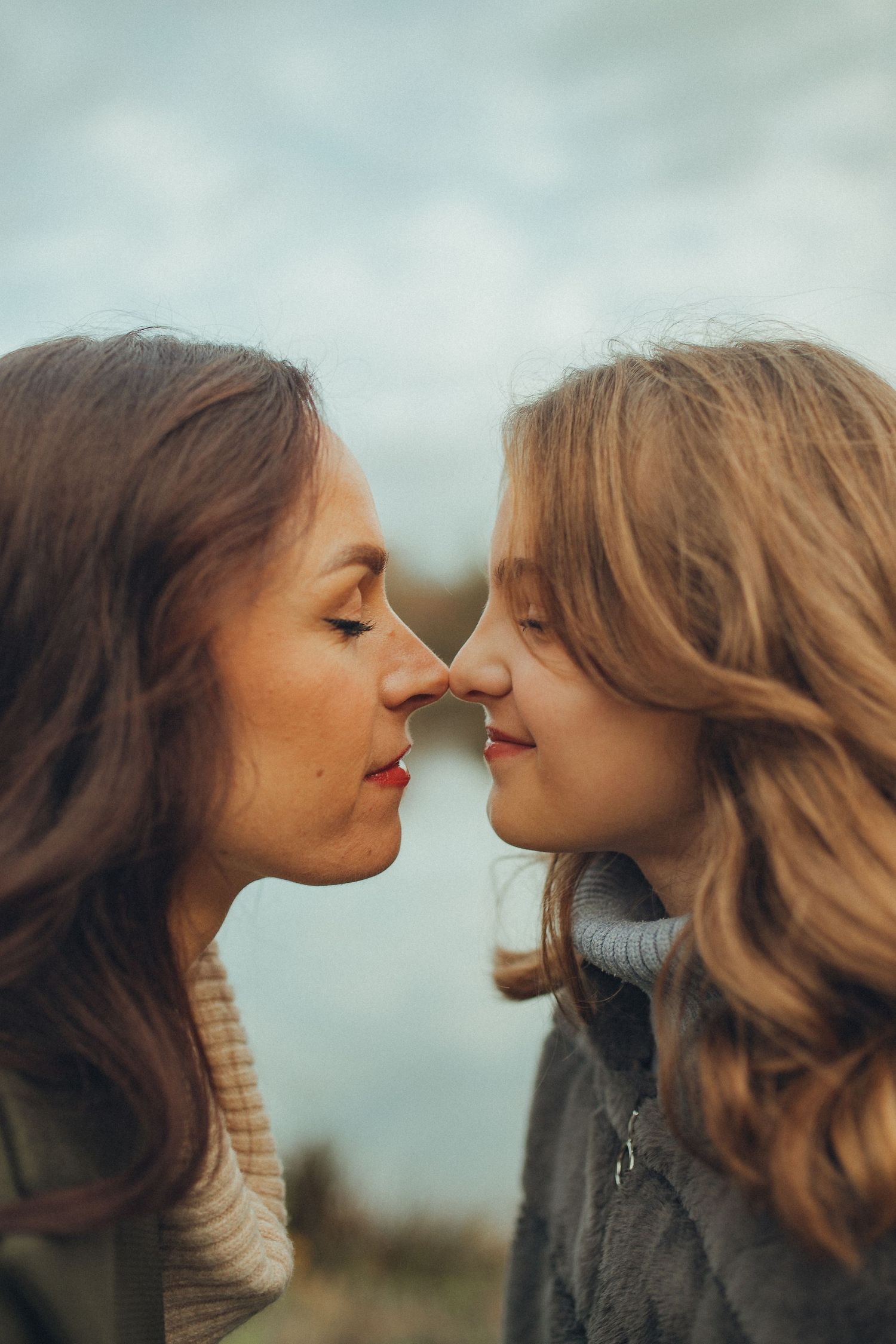 Close intimate portrait of two women touching noses against cloudy sky.