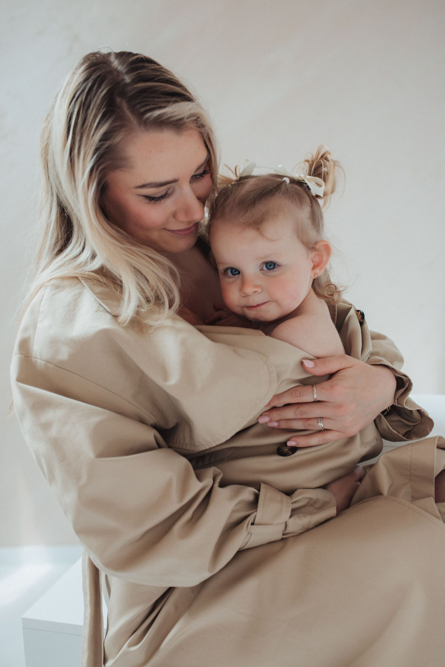 A blonde mother tenderly holds her baby girl in a beige wrap dress against a white background.