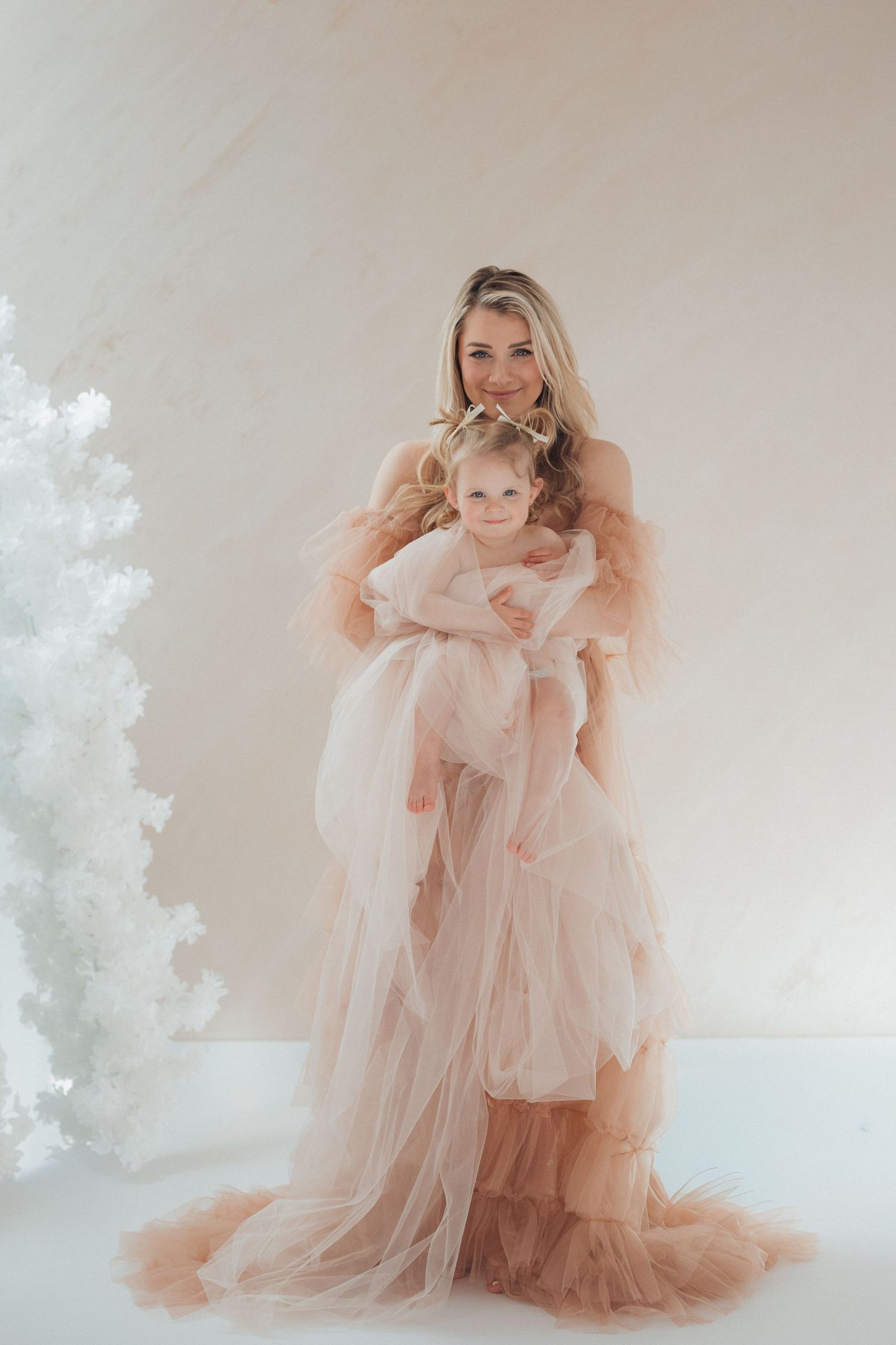 A mother and daughter pose together in matching blush pink tulle gowns beside a white Christmas tree in a studio.