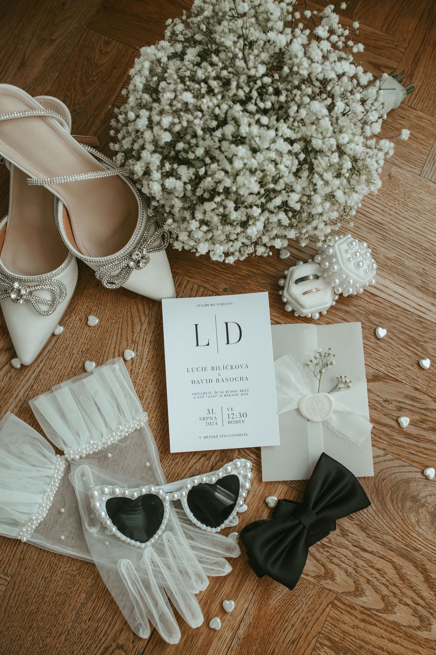 Wedding accessories flatlay with baby's breath bouquet, gloves, heart sunglasses and invitation on wooden floor.