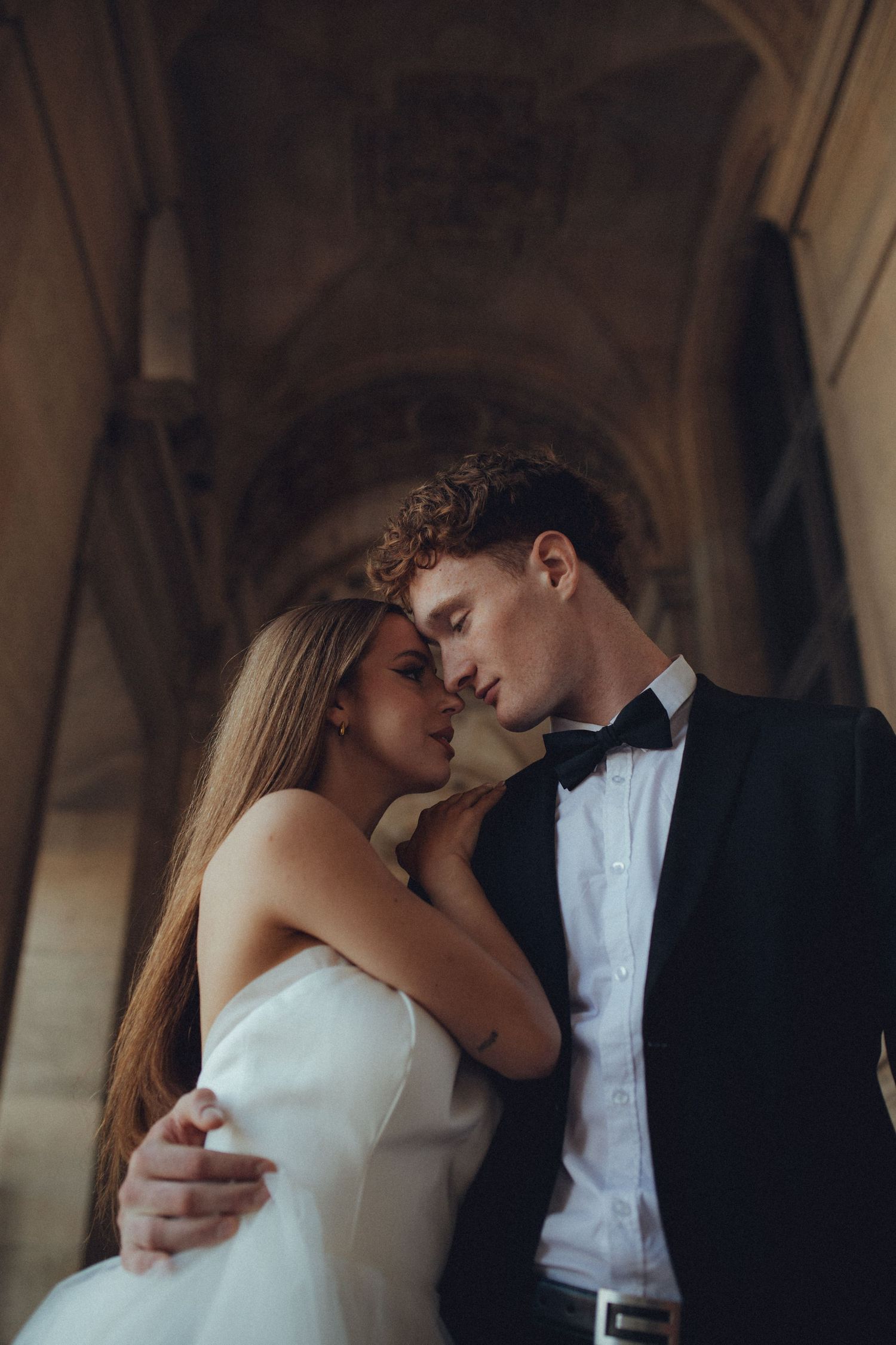 Romantic couple in formal wedding attire embrace intimately under an arched stone hallway in moody lighting.