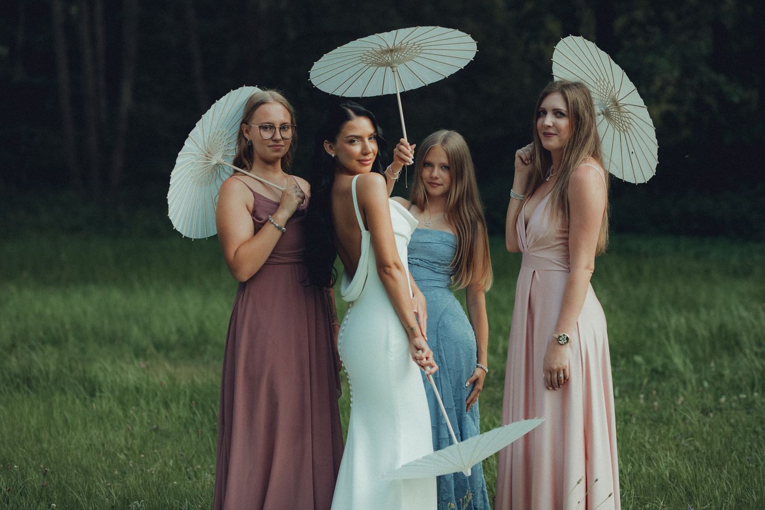 Four women in pastel gowns holding white parasols pose outdoors on green grass near a forest.