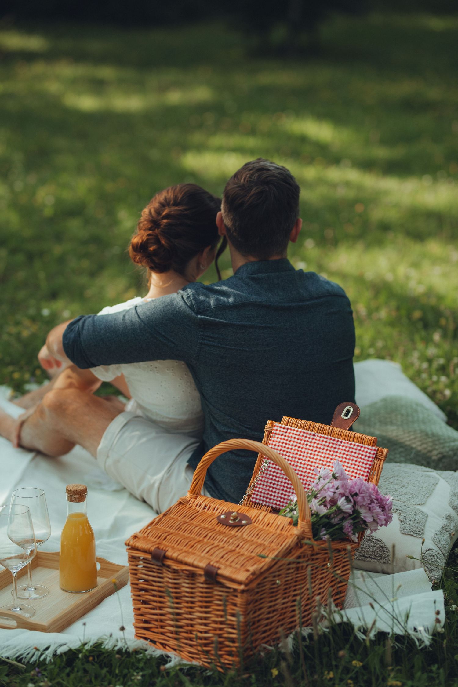 Romantic picnic scene with wicker basket, flowers and drinks on a blanket in sunlit grass.