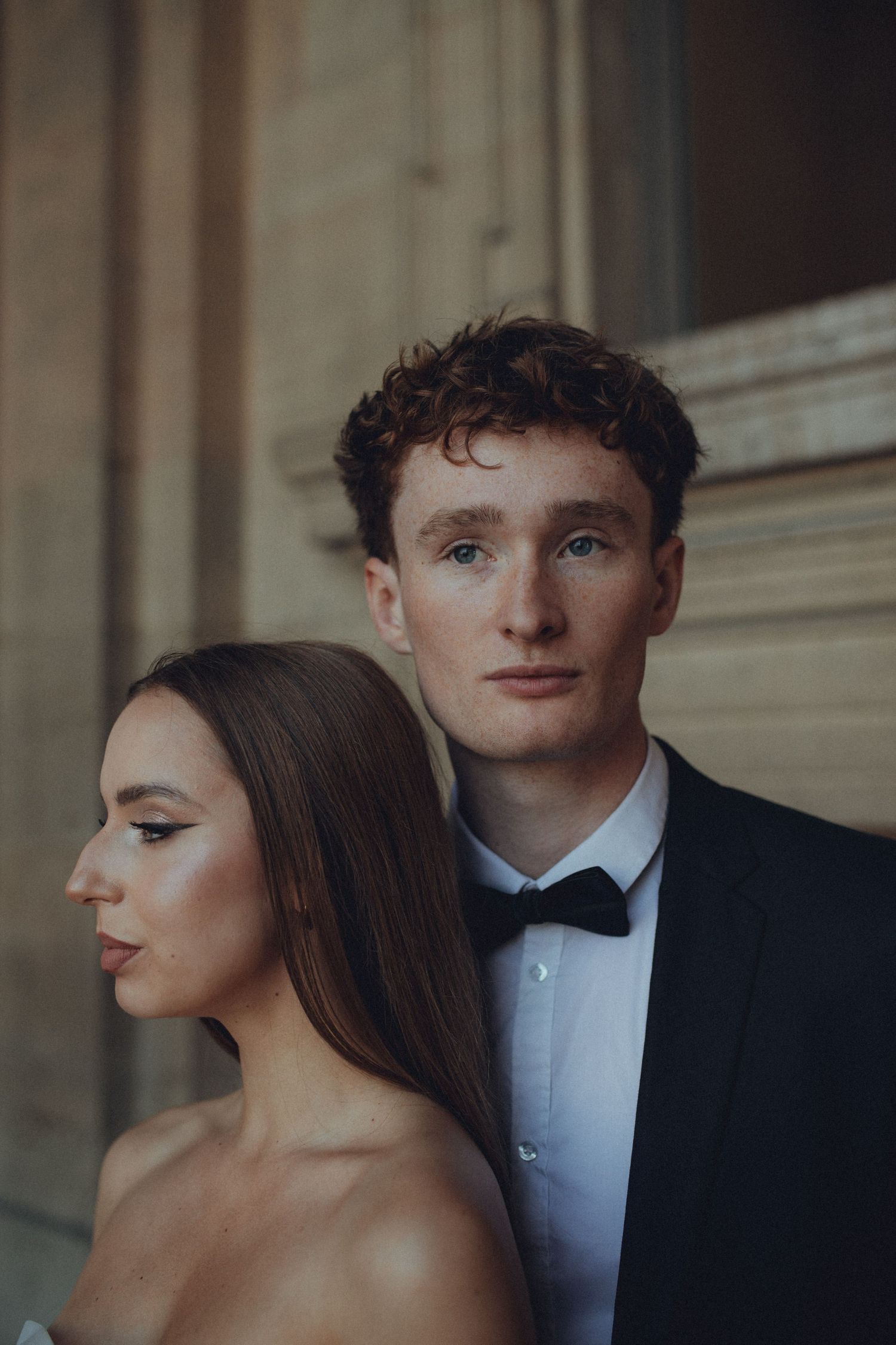 Elegant couple in formal attire pose together at dusk, with him in a black bowtie and her in bare shoulders.