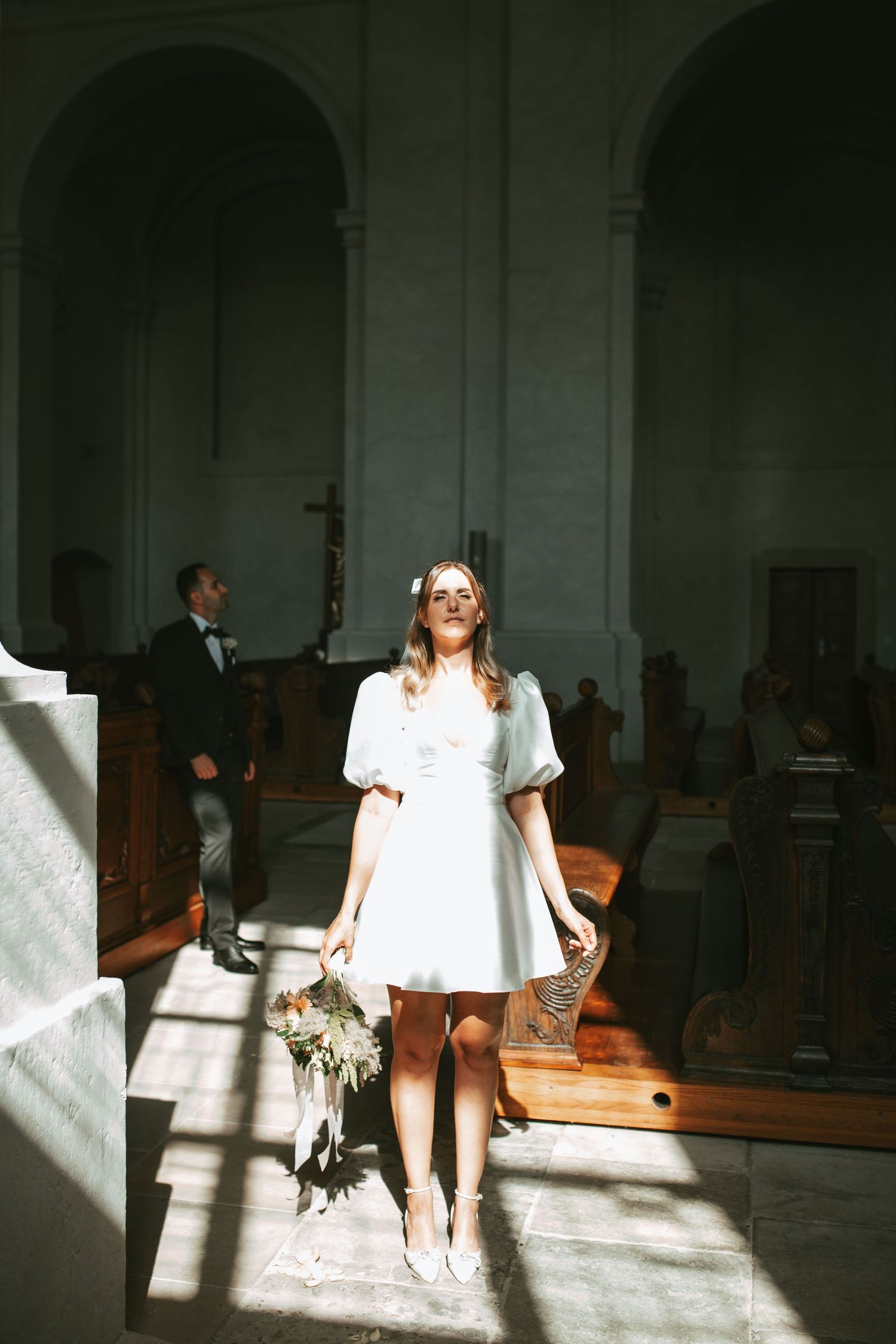 Bride in short white dress standing in sunlit church aisle holding bouquet.