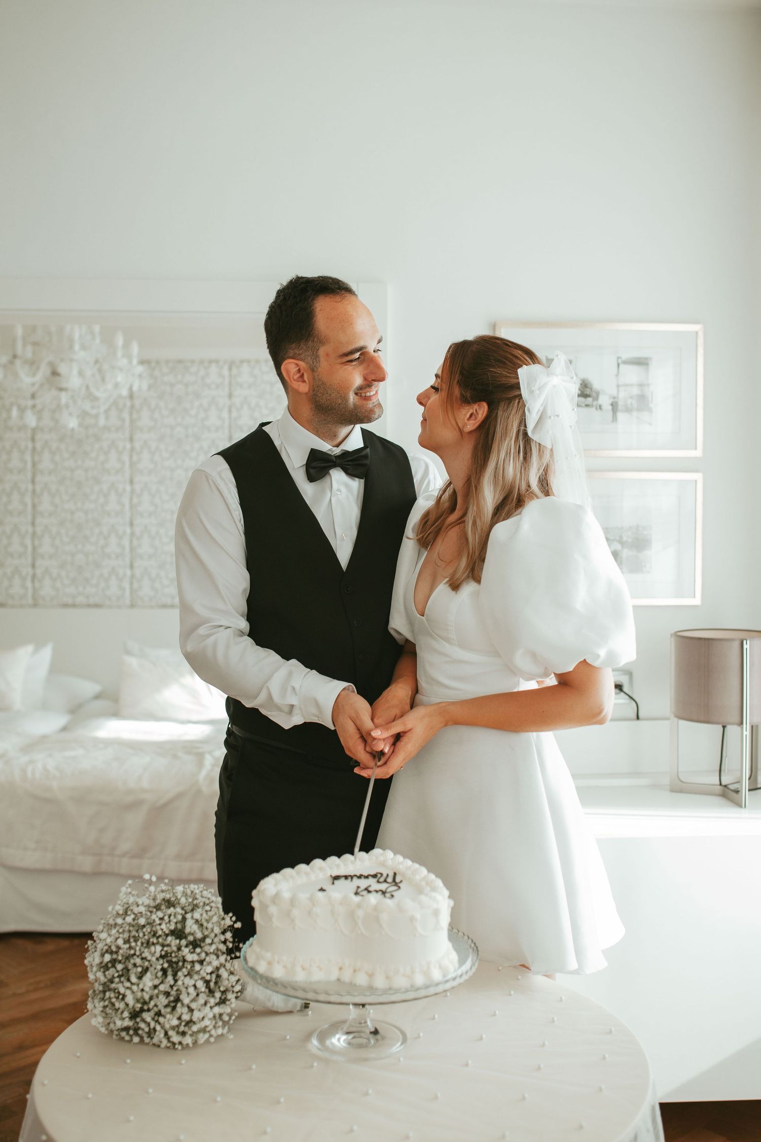 Couple cutting wedding cake with baby's breath flowers in bright modern bedroom setting.