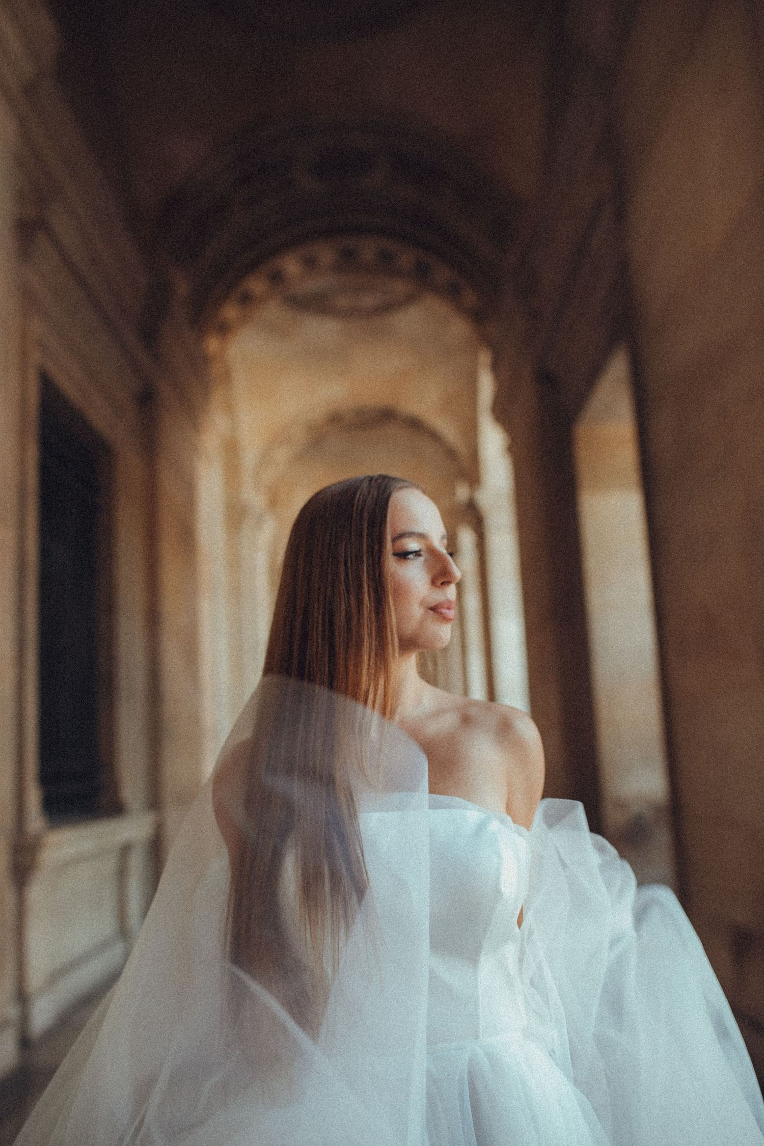 A bride in a white off-shoulder gown and veil poses elegantly in an arched corridor with golden-hour lighting.