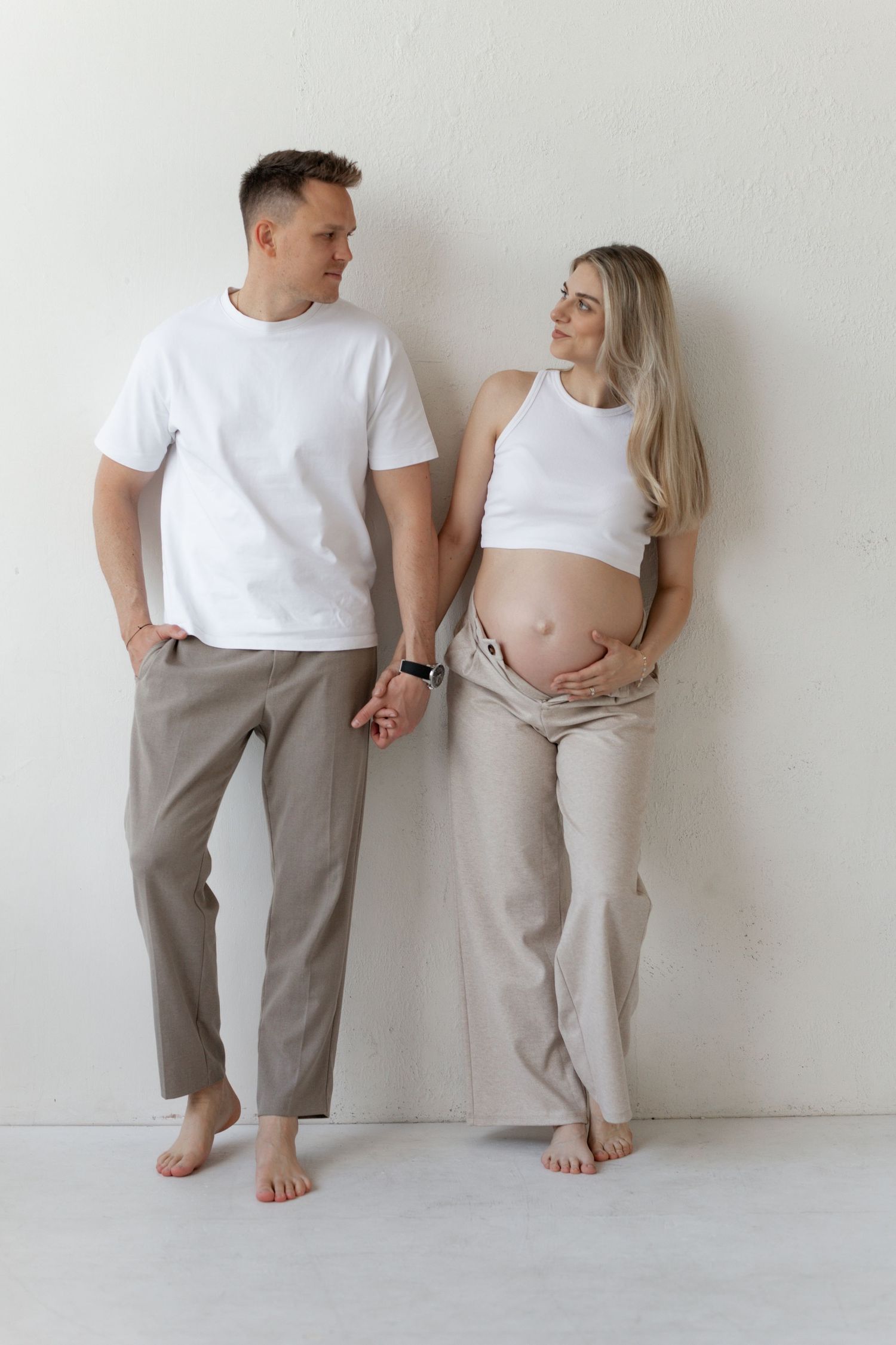 A couple stands against a white wall, the woman visibly pregnant, both wearing neutral tones and barefoot.
