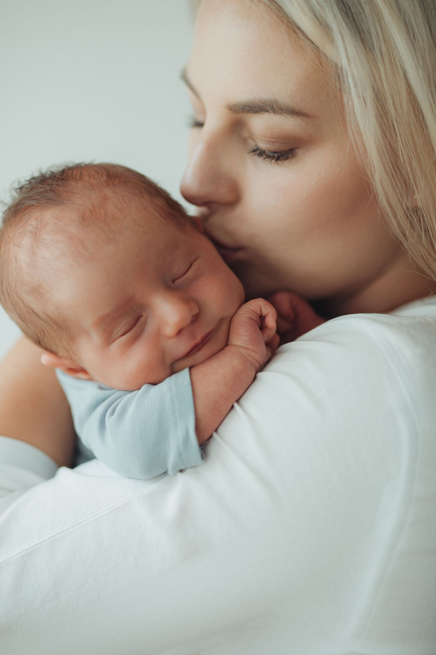 A loving mother gently kisses her sleeping newborn baby dressed in a light blue onesie against a white background.