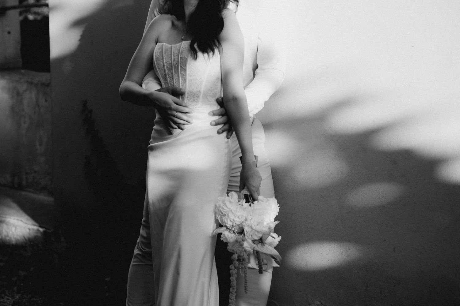 Black and white photo of two women in elegant gowns, one in a wedding dress holding a bouquet, embracing outdoors.