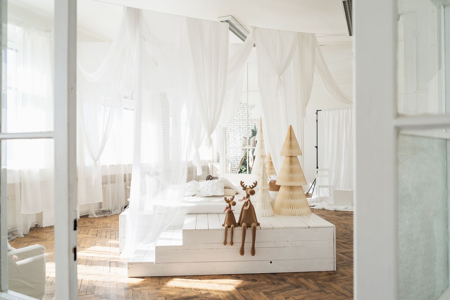 Bright white minimalist bedroom with sheer curtains, wooden floor, and decorative reindeer figurines on a raised platform.