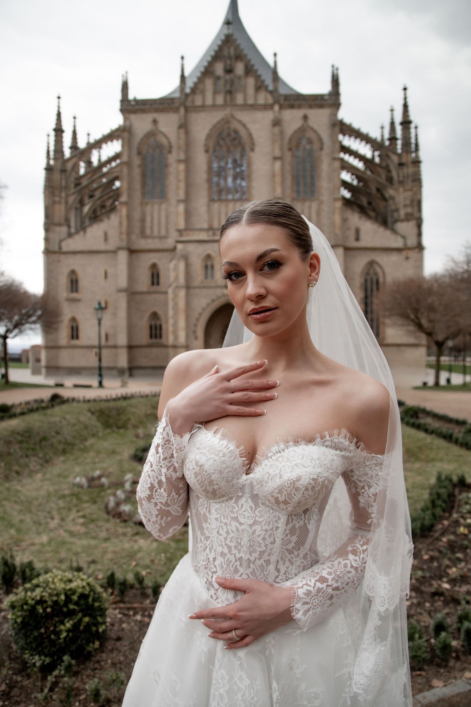 Bride in lace off-shoulder gown and veil poses before Gothic cathedral in garden setting.