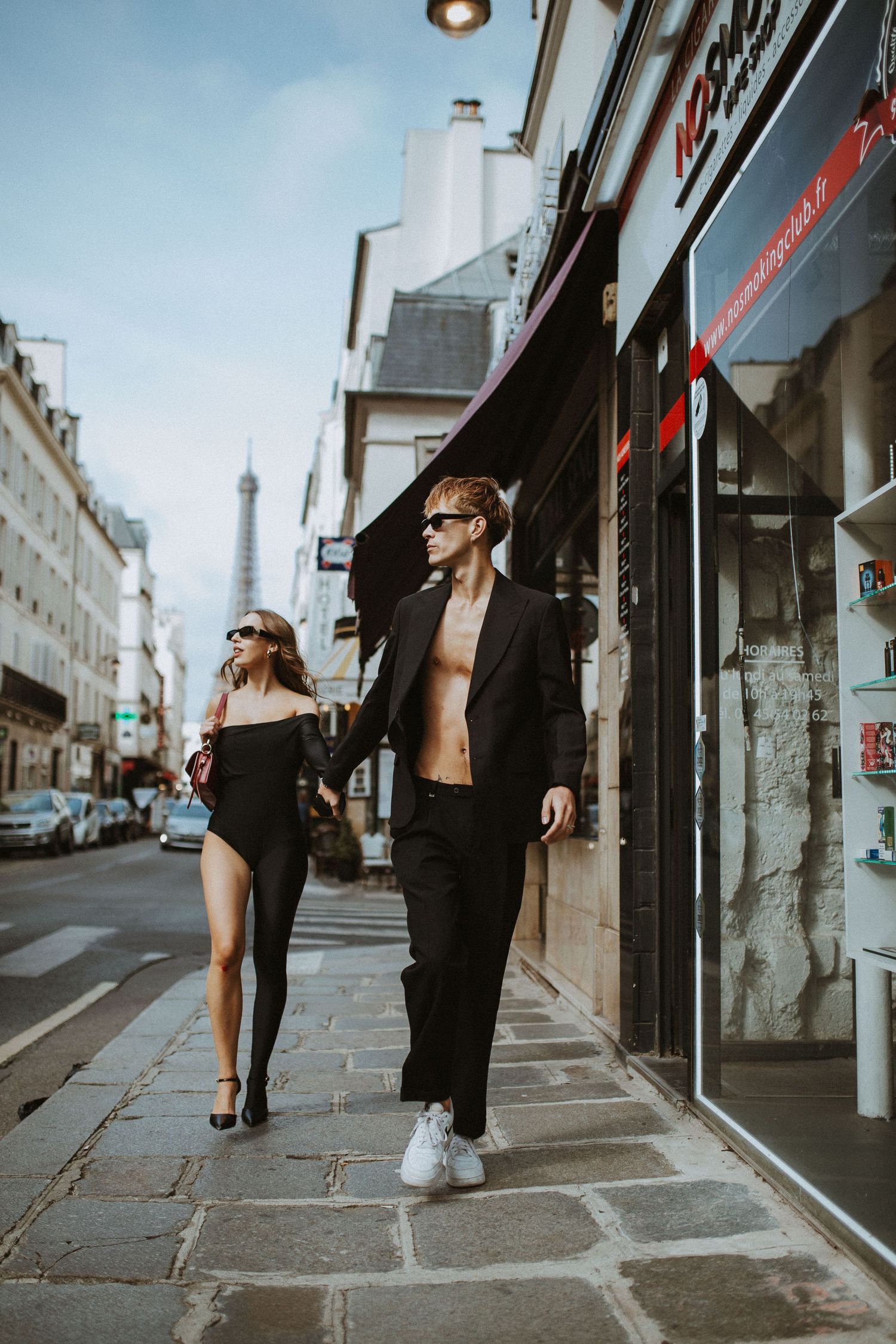 A stylish couple walks down a Parisian street in black outfits with sunglasses on a sunny afternoon.