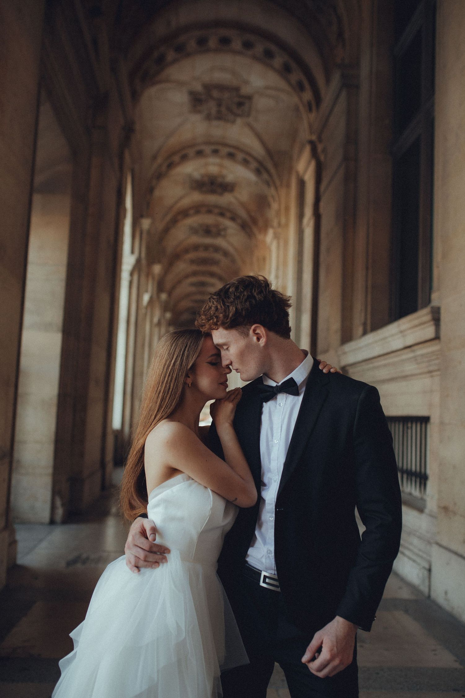 A romantic couple in wedding attire stands close together in an ornate arched corridor.