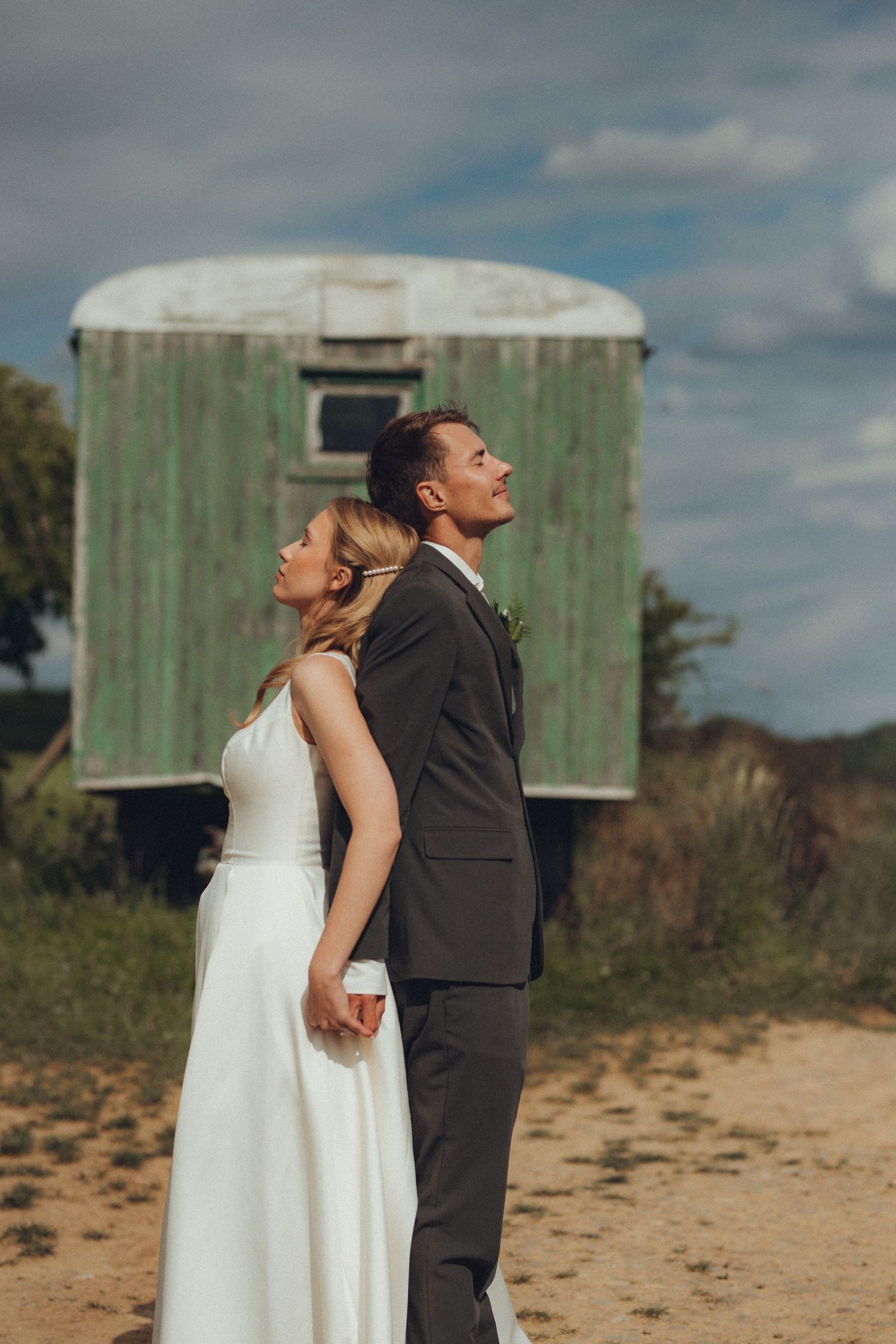 Wedding couple poses back-to-back against rustic green backdrop outdoors.