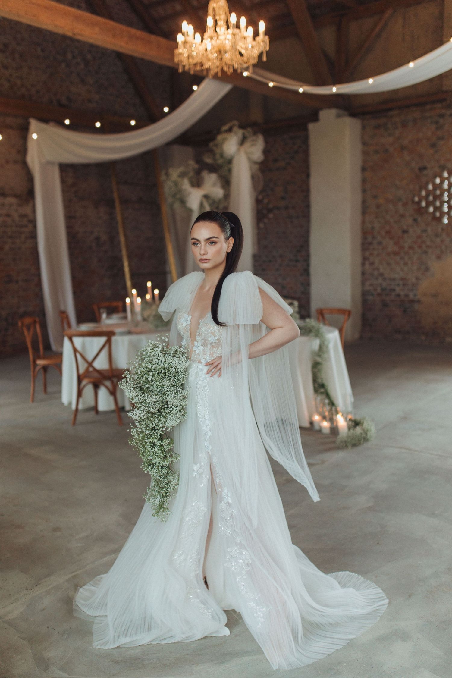 Elegant bride in flowing white lace gown and sheer cape poses in rustic venue with chandelier and draped fabric.