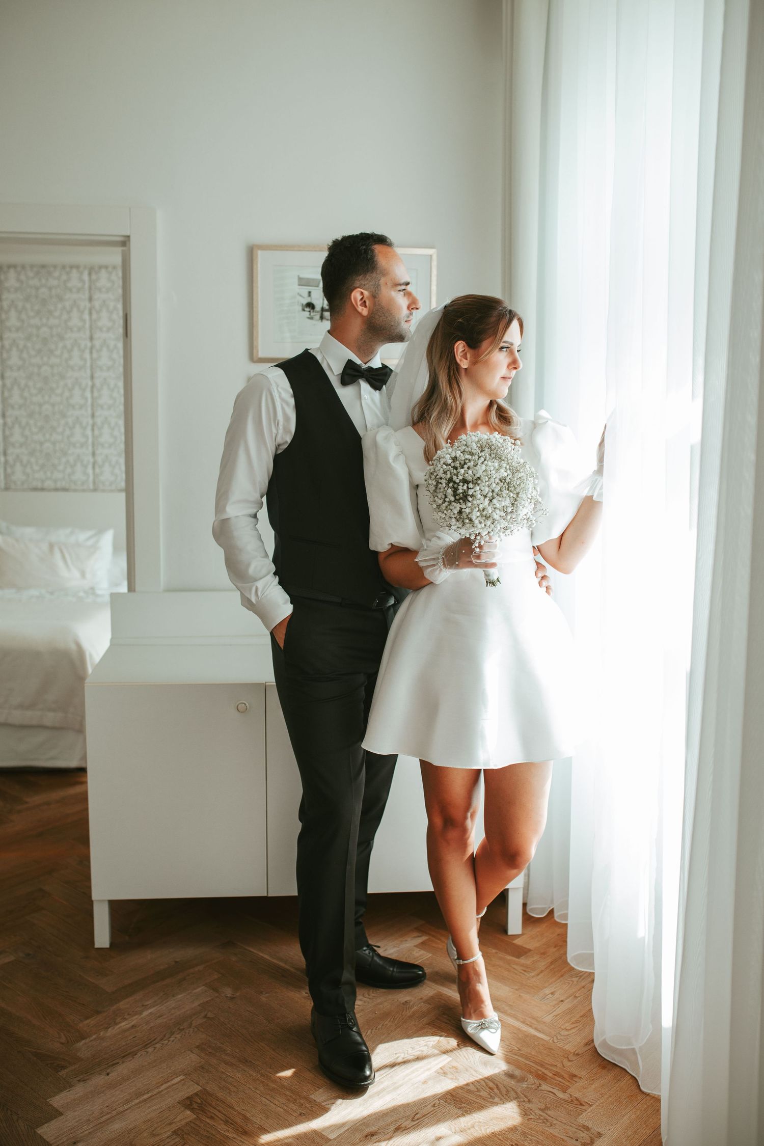 Wedding couple standing by bright window in modern bedroom with herringbone floors.
