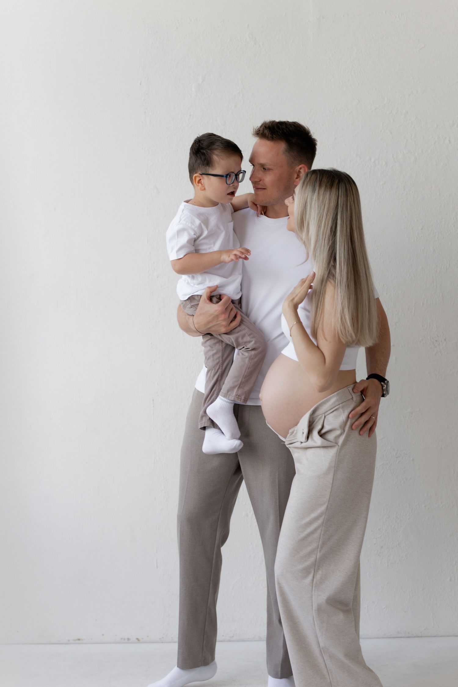 Happy family portrait with pregnant woman, man holding toddler in glasses, posing together against white wall.