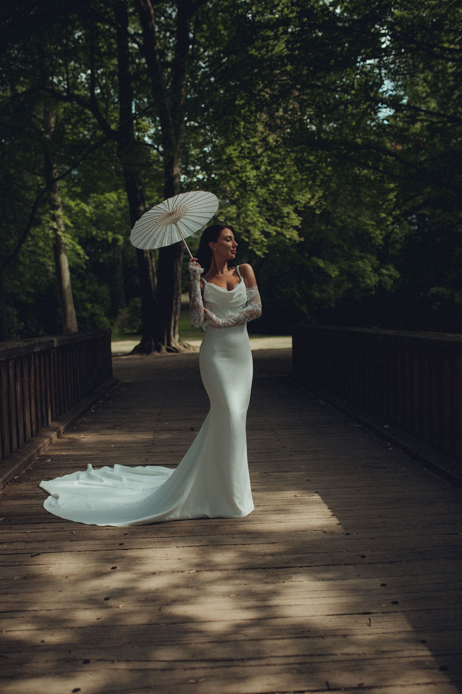 Bride in white mermaid gown with train holds parasol on wooden bridge surrounded by lush green trees.