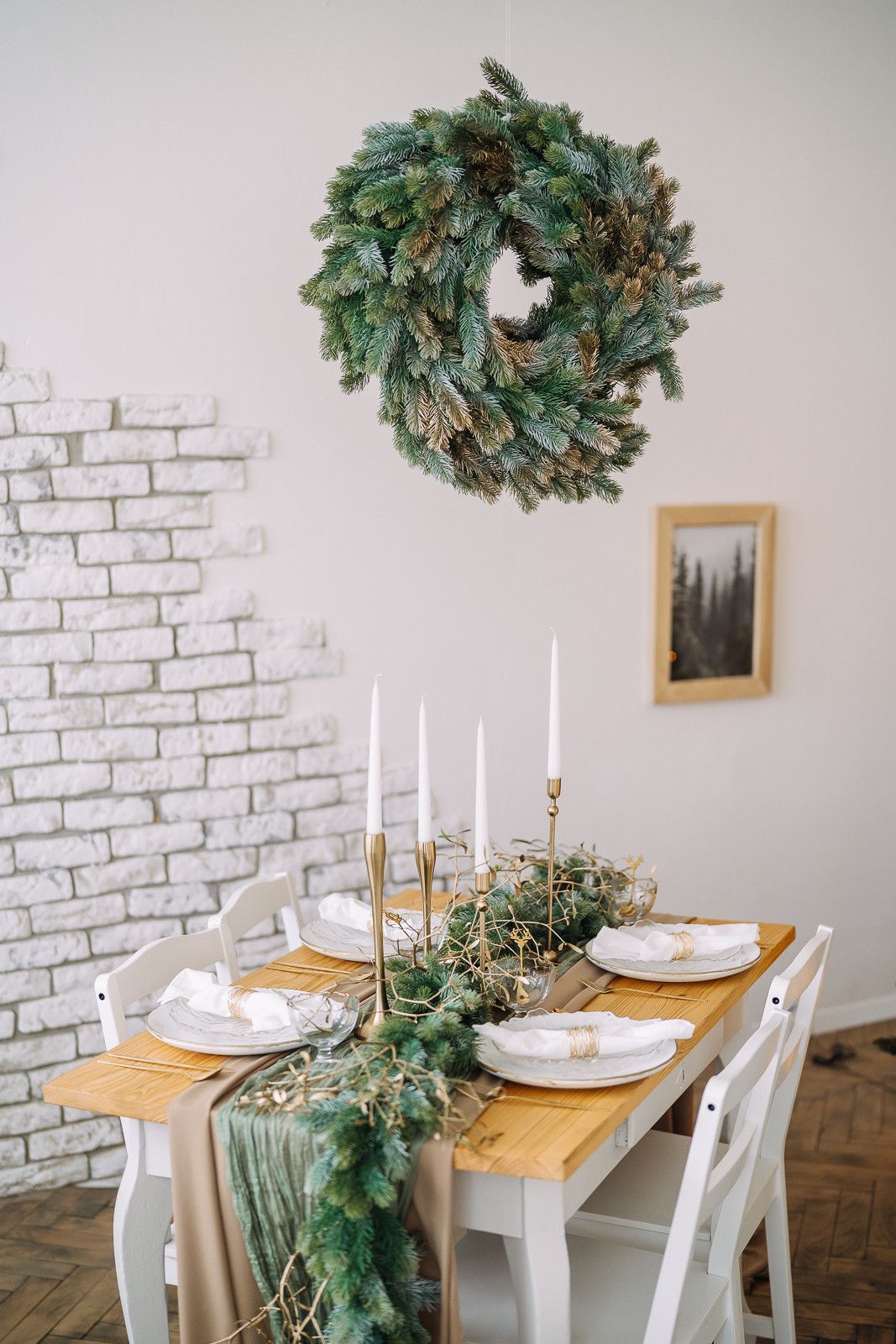 Rustic dining setup with white chairs, wood table, brick wall accent, and evergreen wreath decor.