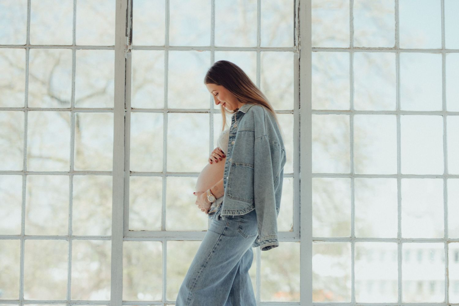 Woman in casual denim outfit standing by bright window in minimal interior setting.