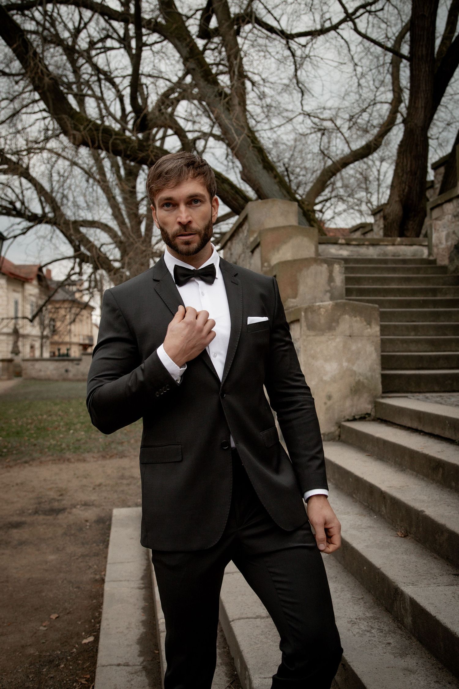 Elegant man in black tuxedo with bow tie adjusting collar near stone steps and bare trees outdoors.