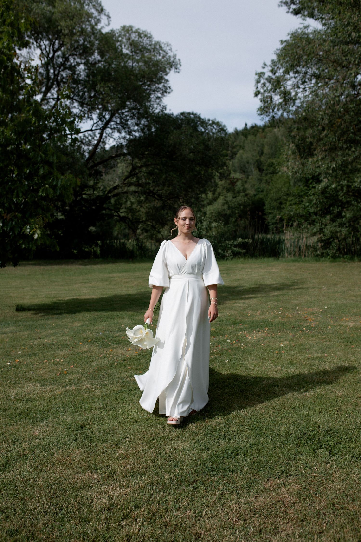 Bride in white flowing dress with flutter sleeves walks across green lawn holding bouquet, surrounded by lush trees.