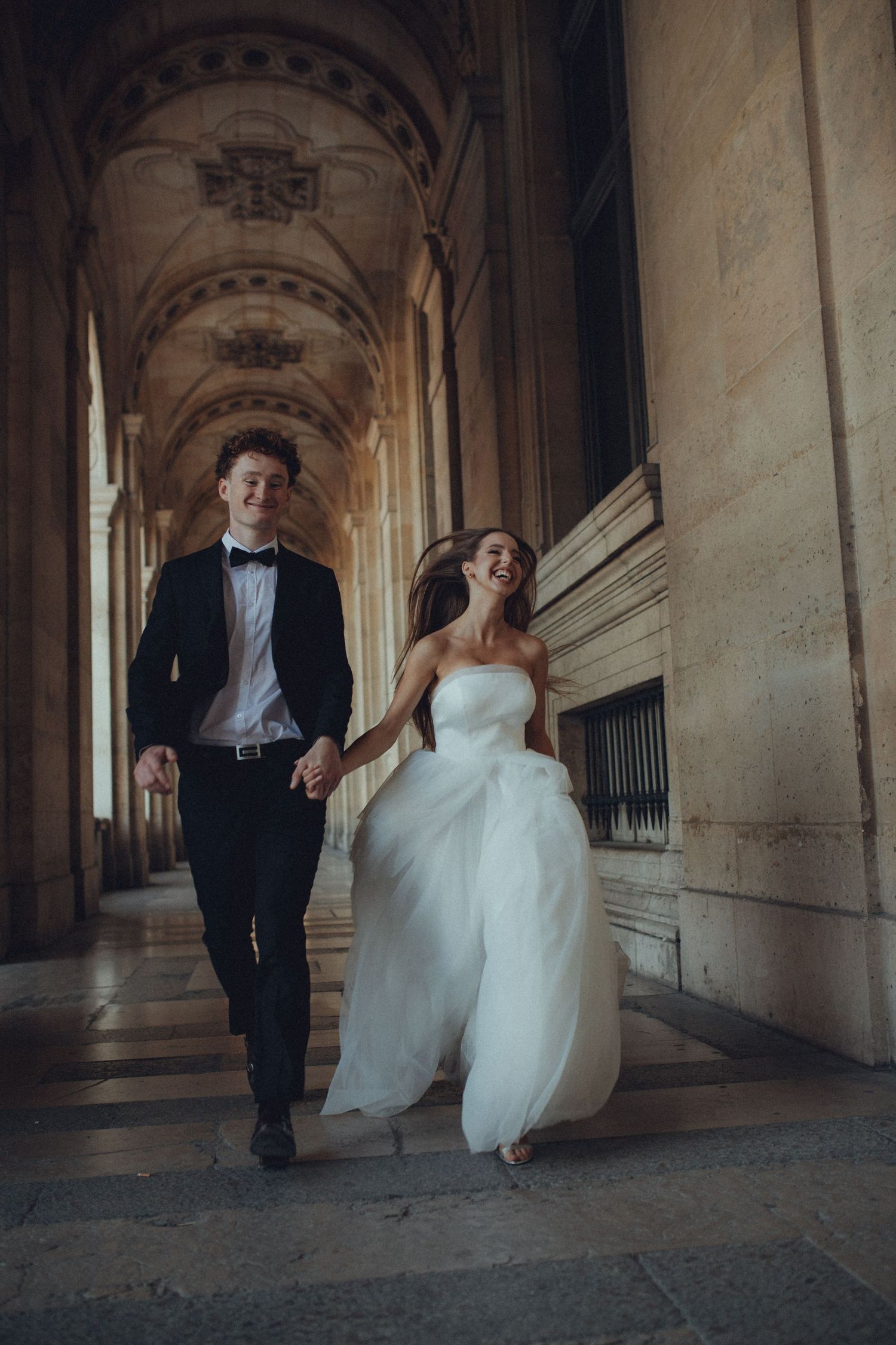A couple in wedding attire runs playfully through an elegant stone archway corridor.