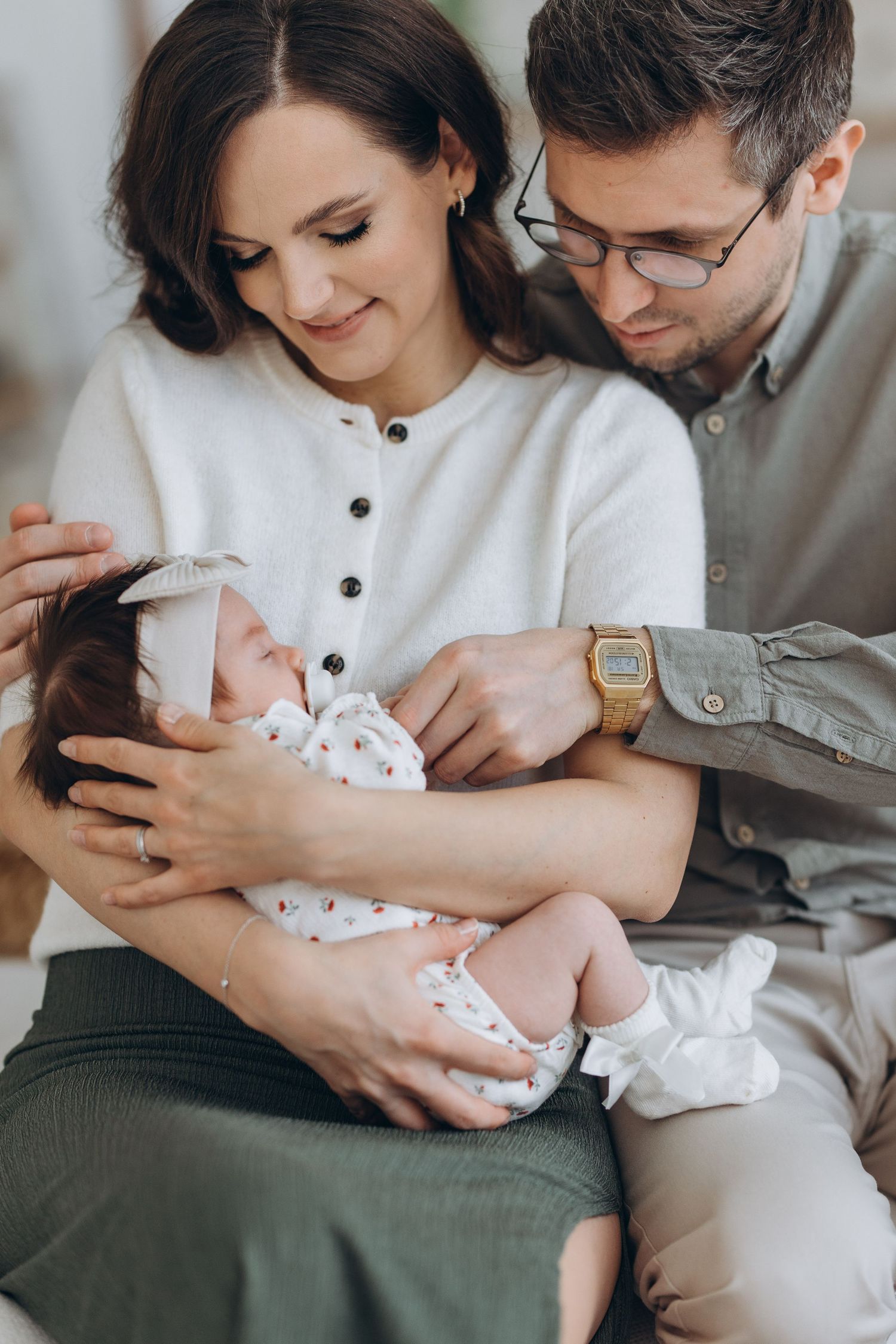 Couple holding newborn baby in intimate indoor photoshoot with soft lighting.