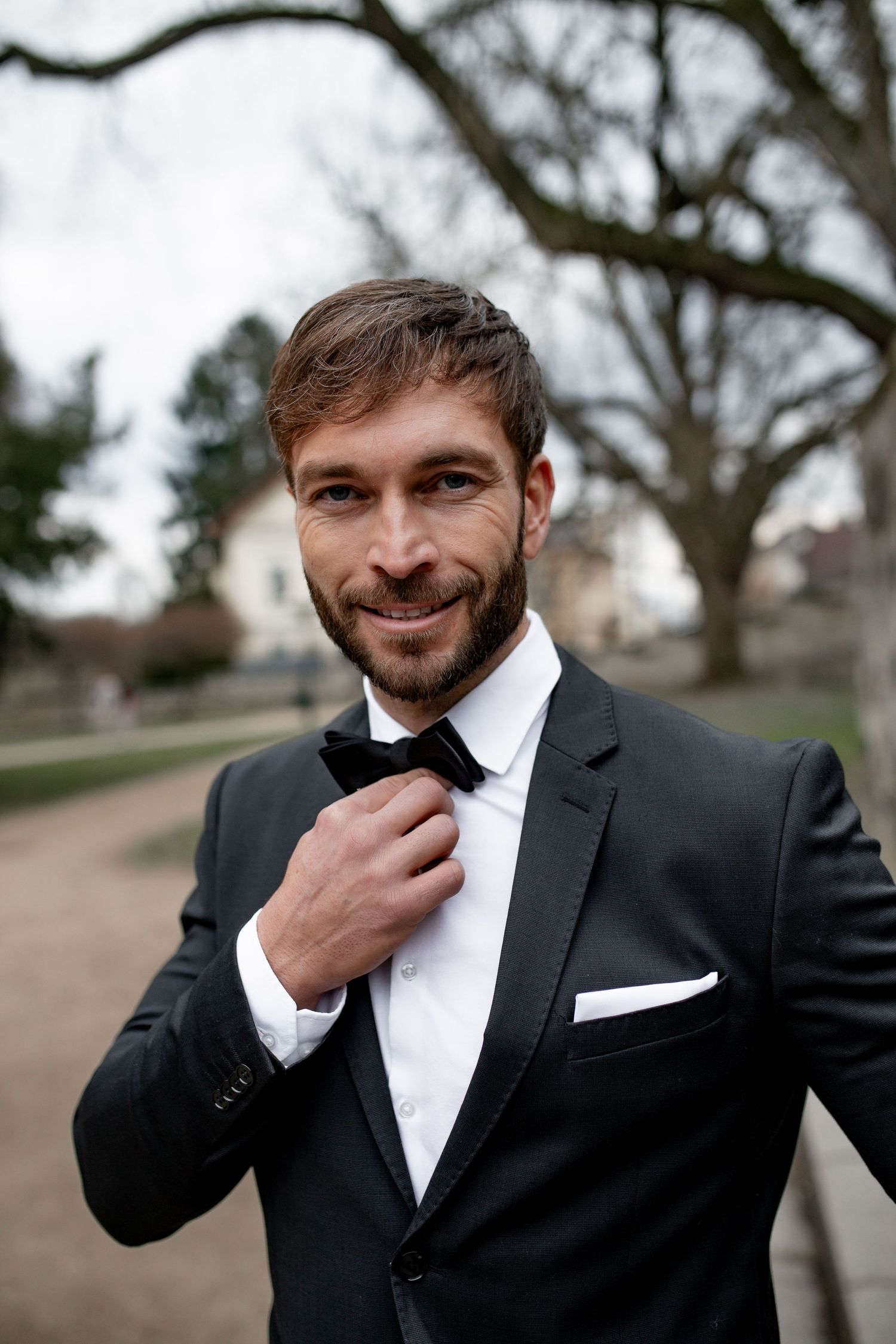 Smiling bearded man in black tuxedo adjusting bow tie outdoors in a park setting.