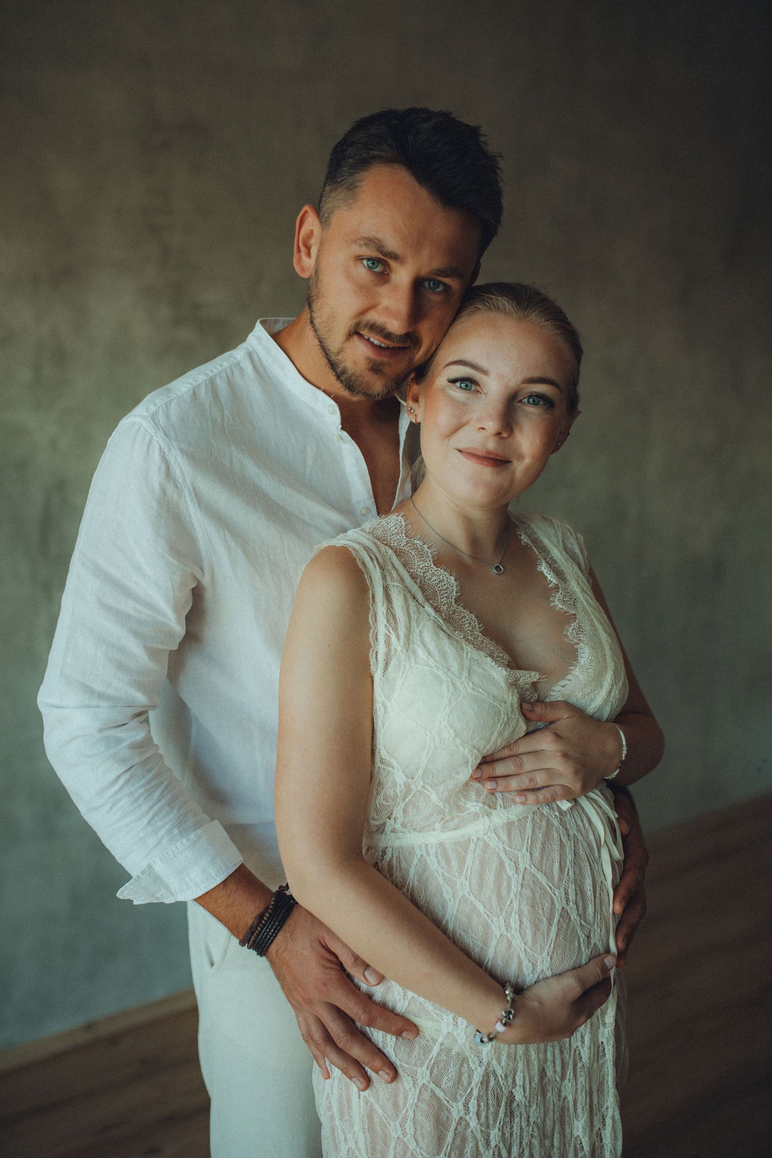 Intimate maternity photoshoot showing couple embracing in white clothing against textured grey wall.