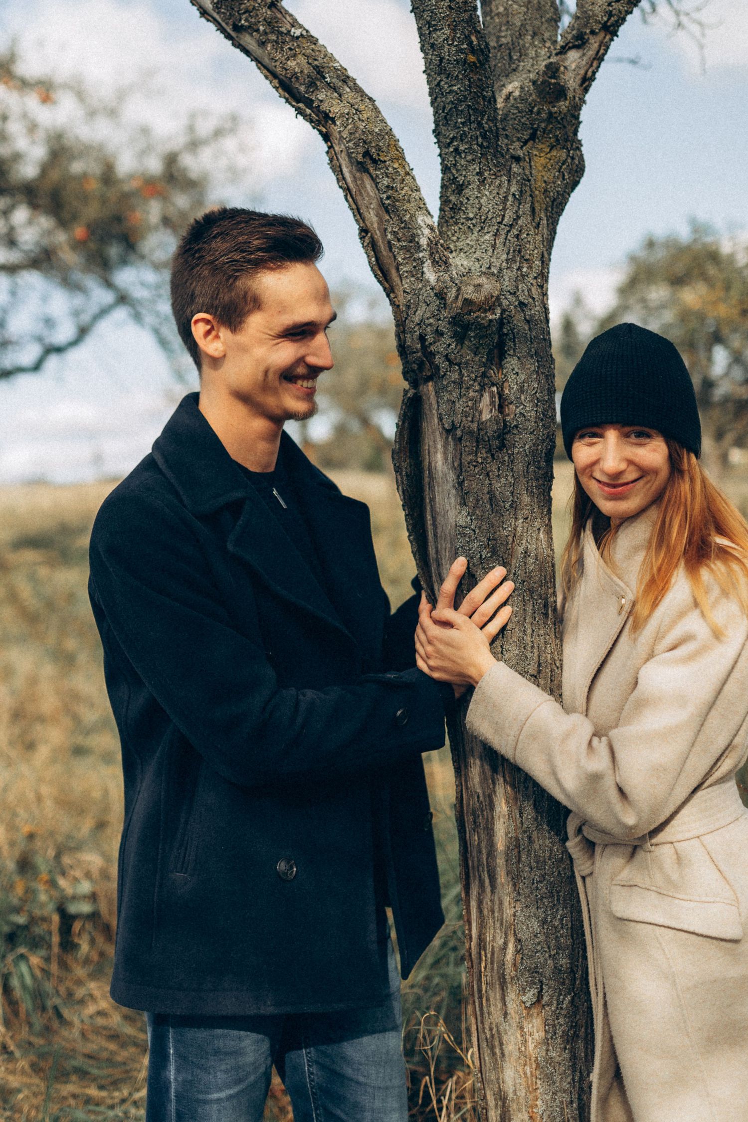 A couple shares a joyful moment together while leaning against a tree trunk in an autumn field.