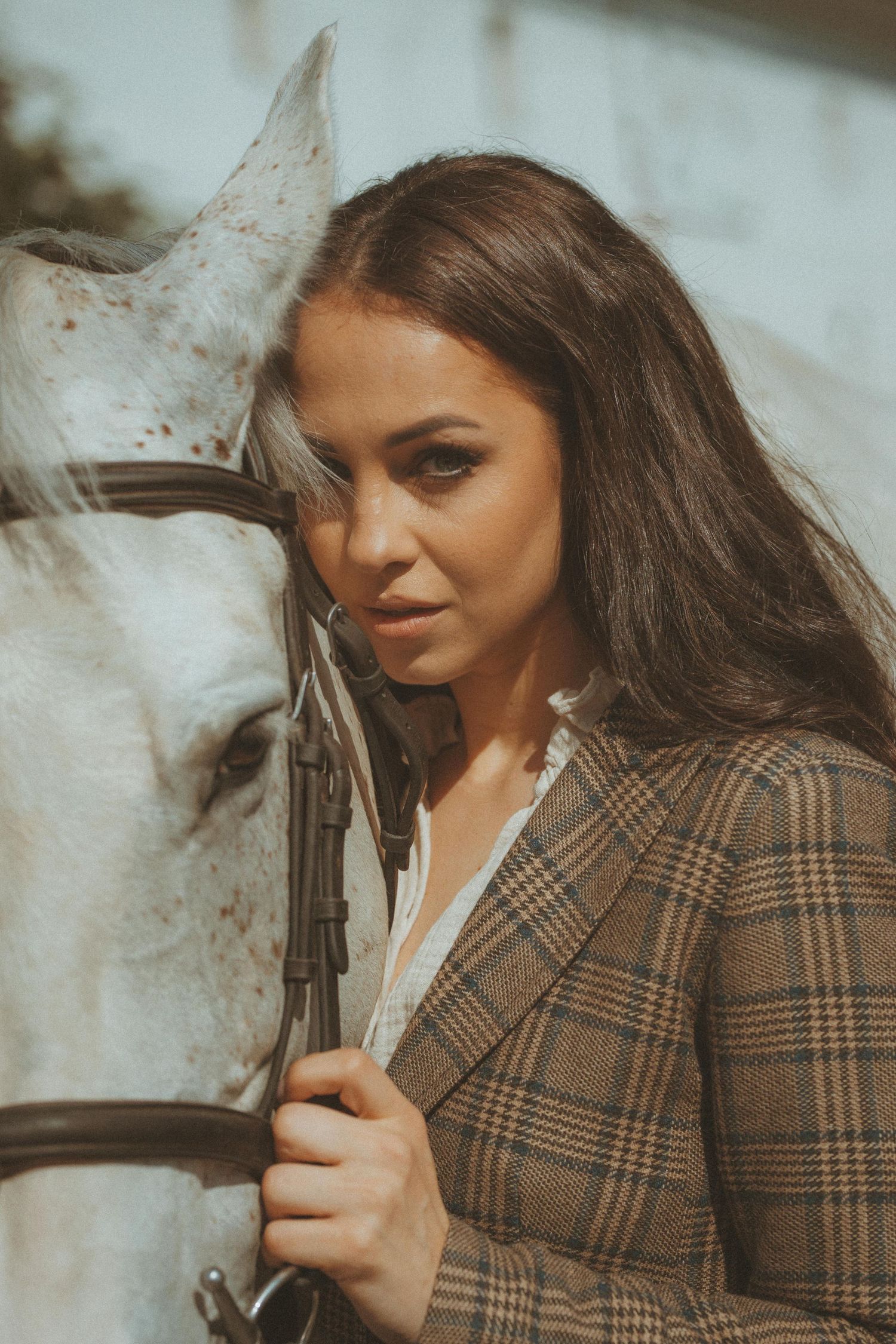 A woman in a plaid blazer poses next to a white horse in a warm-toned outdoor portrait.