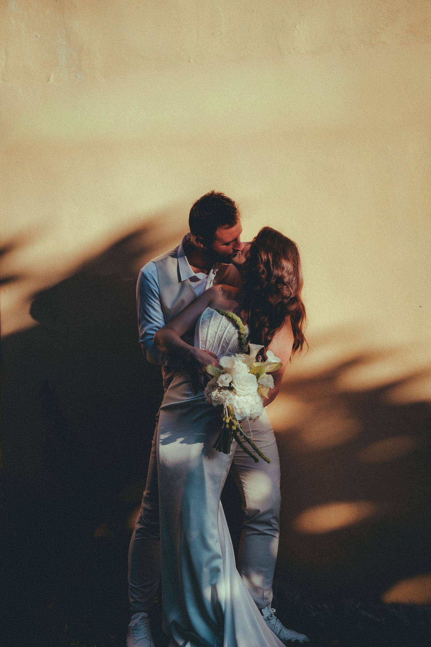 Bride and groom kissing against a warm golden wall, bride holding white floral bouquet in elegant satin wedding dress.