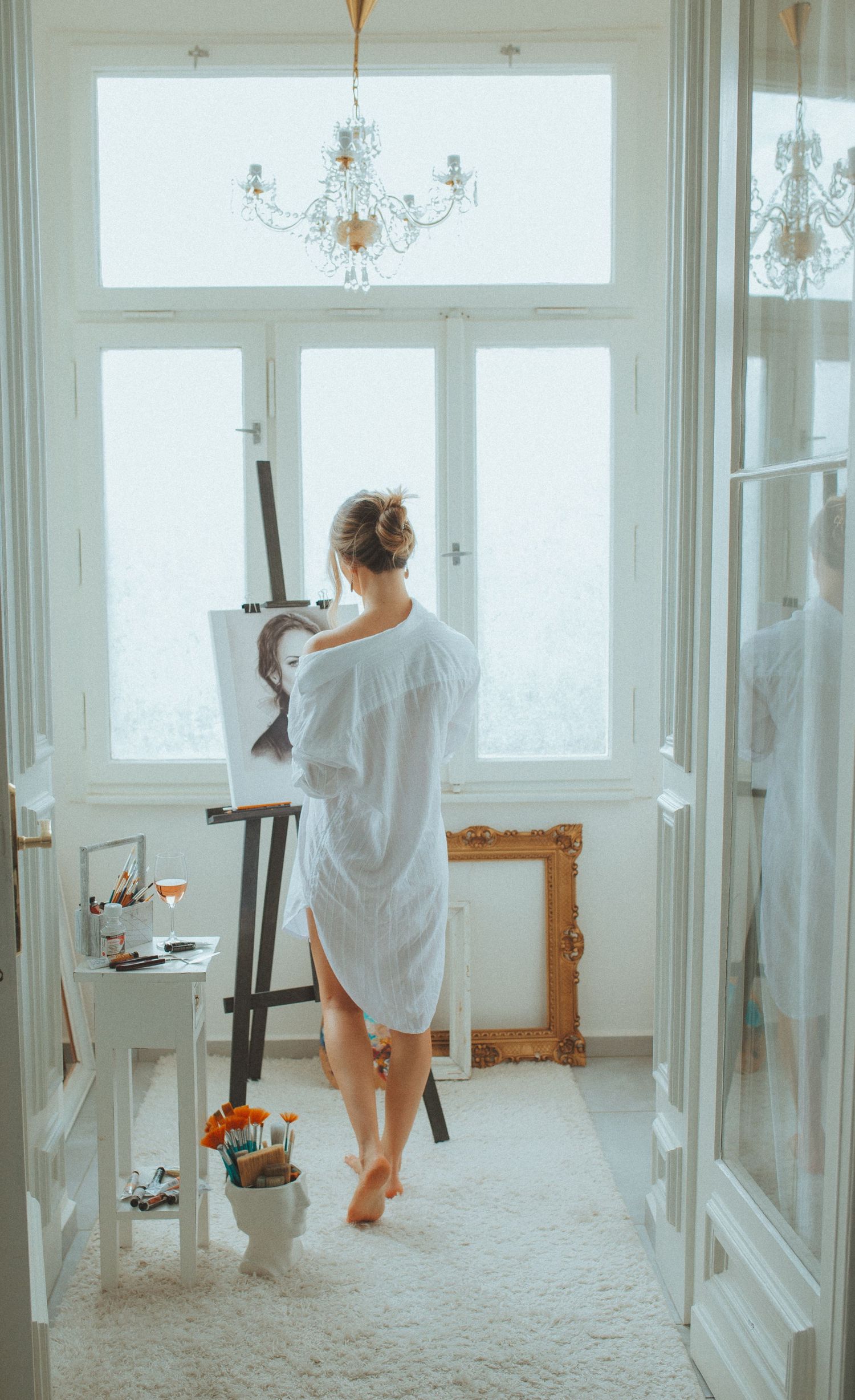 Artist in white dress working in bright sunlit studio space with elegant chandeliers and windows.
